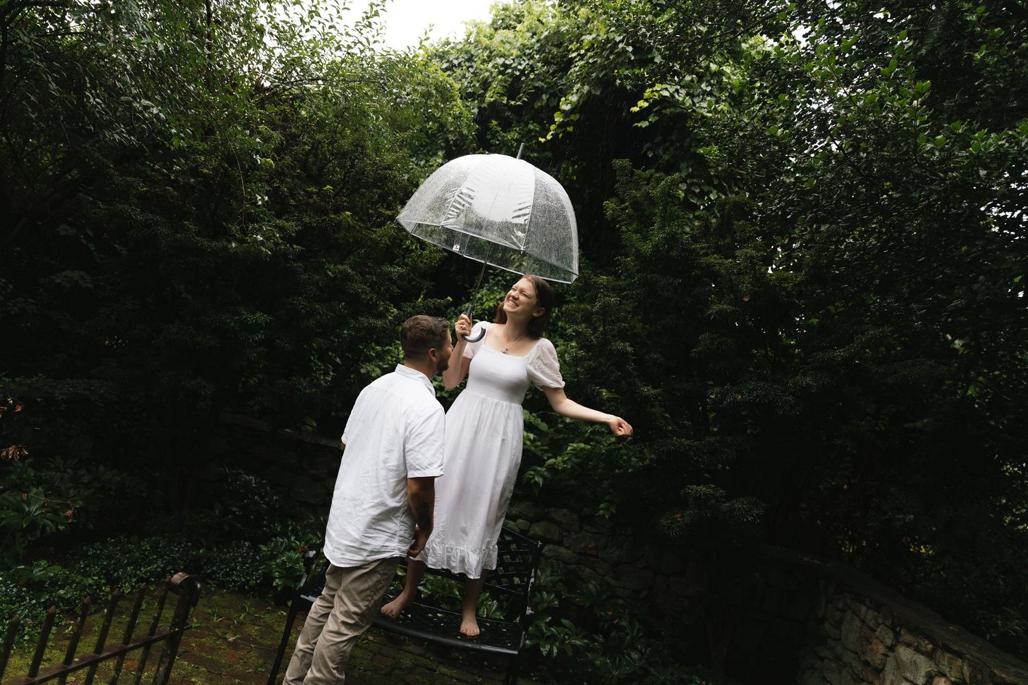 Couple shares romantic moment under clear umbrella in lush garden setting.