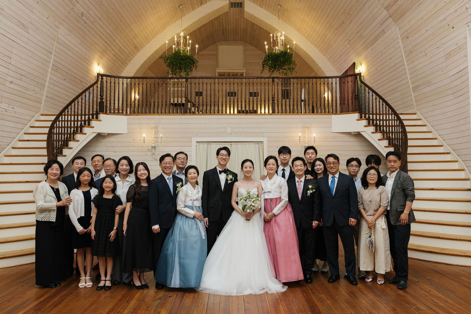 Wedding group photo with bride and groom surrounded by family in elegant wood chapel with dual staircases.