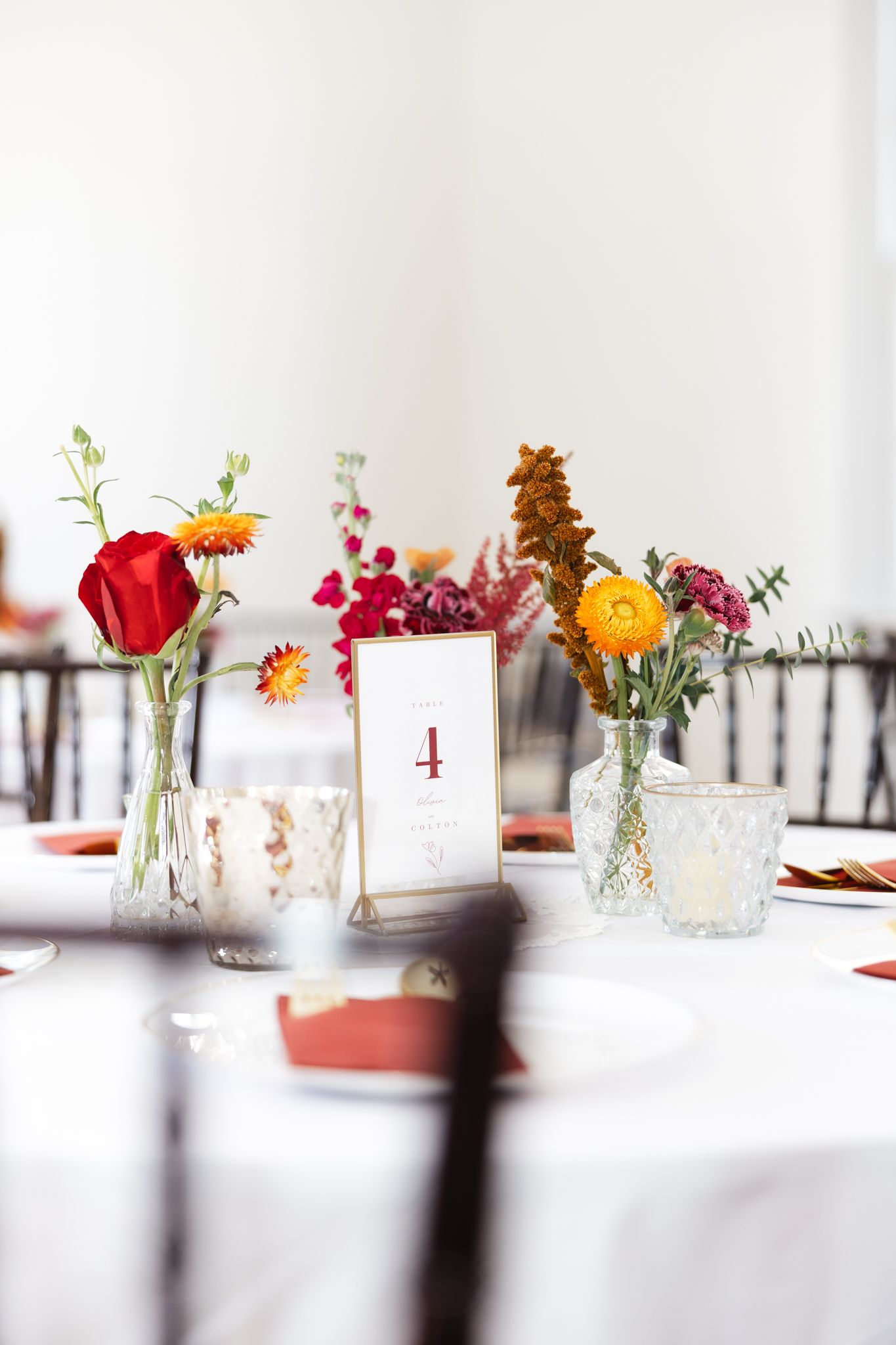 Colorful wedding table centerpiece with red, orange, and pink flowers in glass vases.