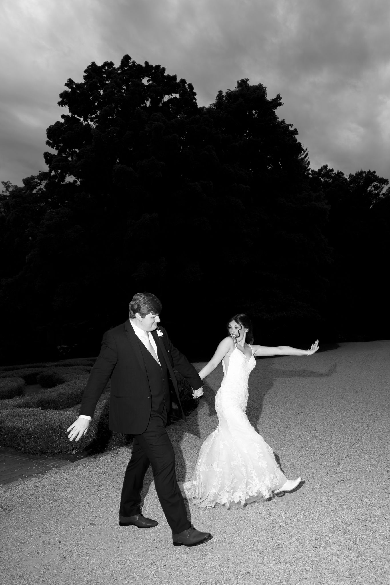 Black and white photo of a bride and groom holding hands, walking on a gravel path with dramatic cloudy sky.