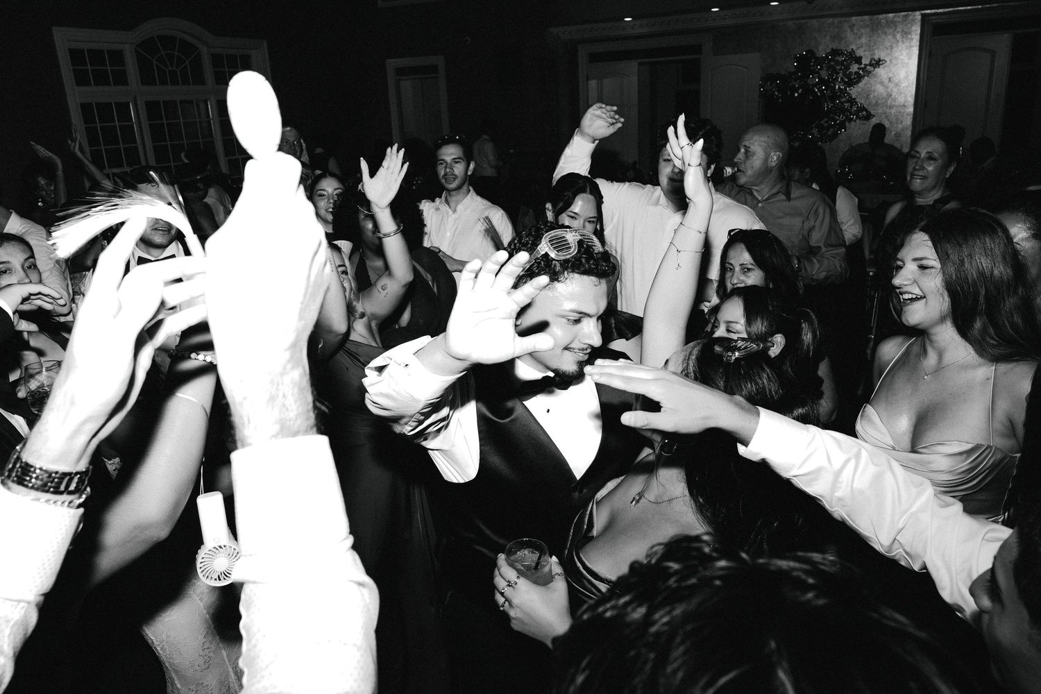 Black and white photo of a groom in tuxedo surrounded by celebrating wedding guests on a lively dance floor.