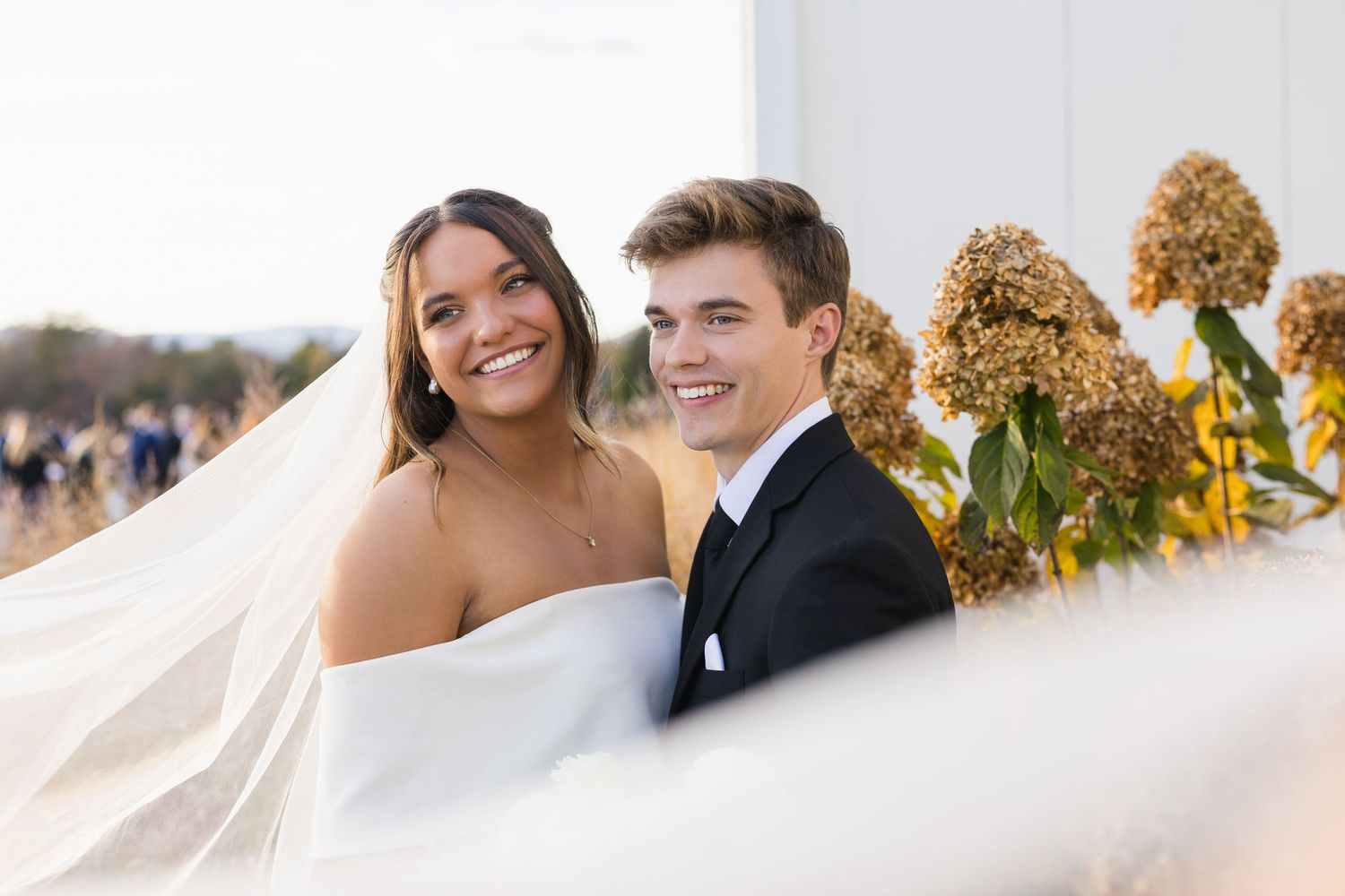 A smiling bride and groom pose together outdoors with dried hydrangea flowers in the background.