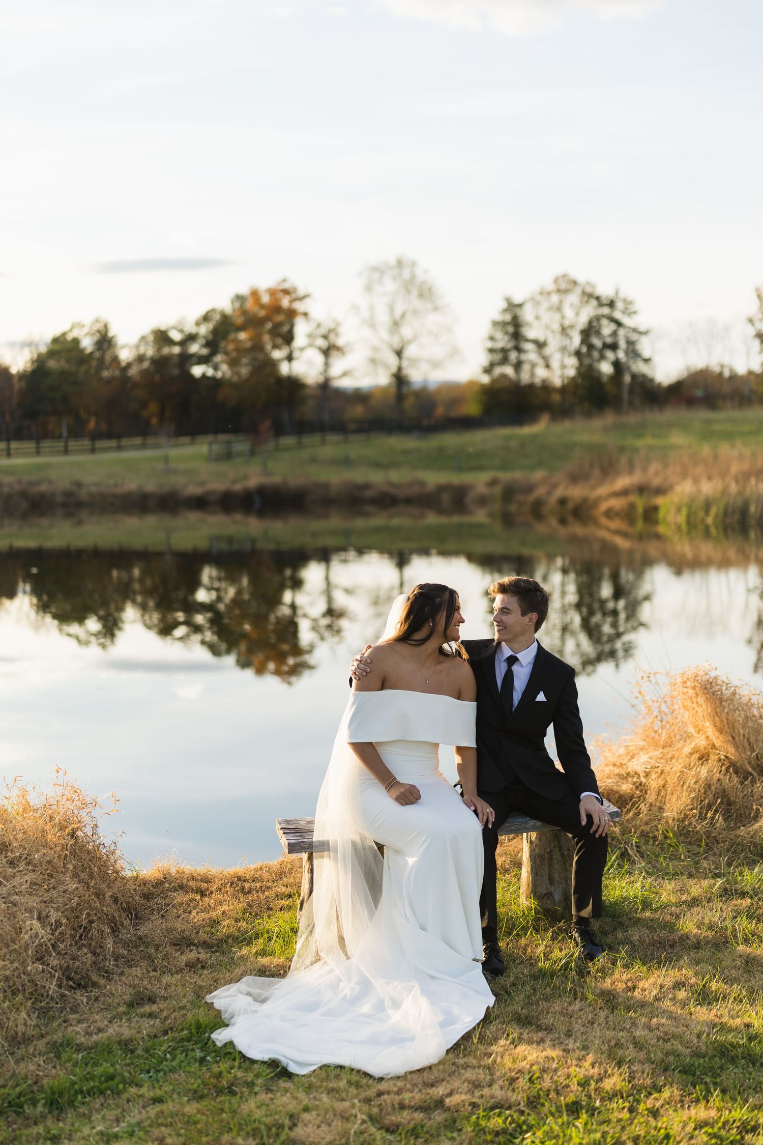 Bride and groom sit together by a serene reflective pond surrounded by autumn foliage on their wedding day.