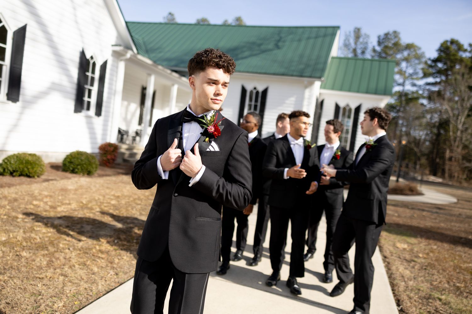 Groom in black tuxedo with boutonniere posing outside white chapel with groomsmen behind.