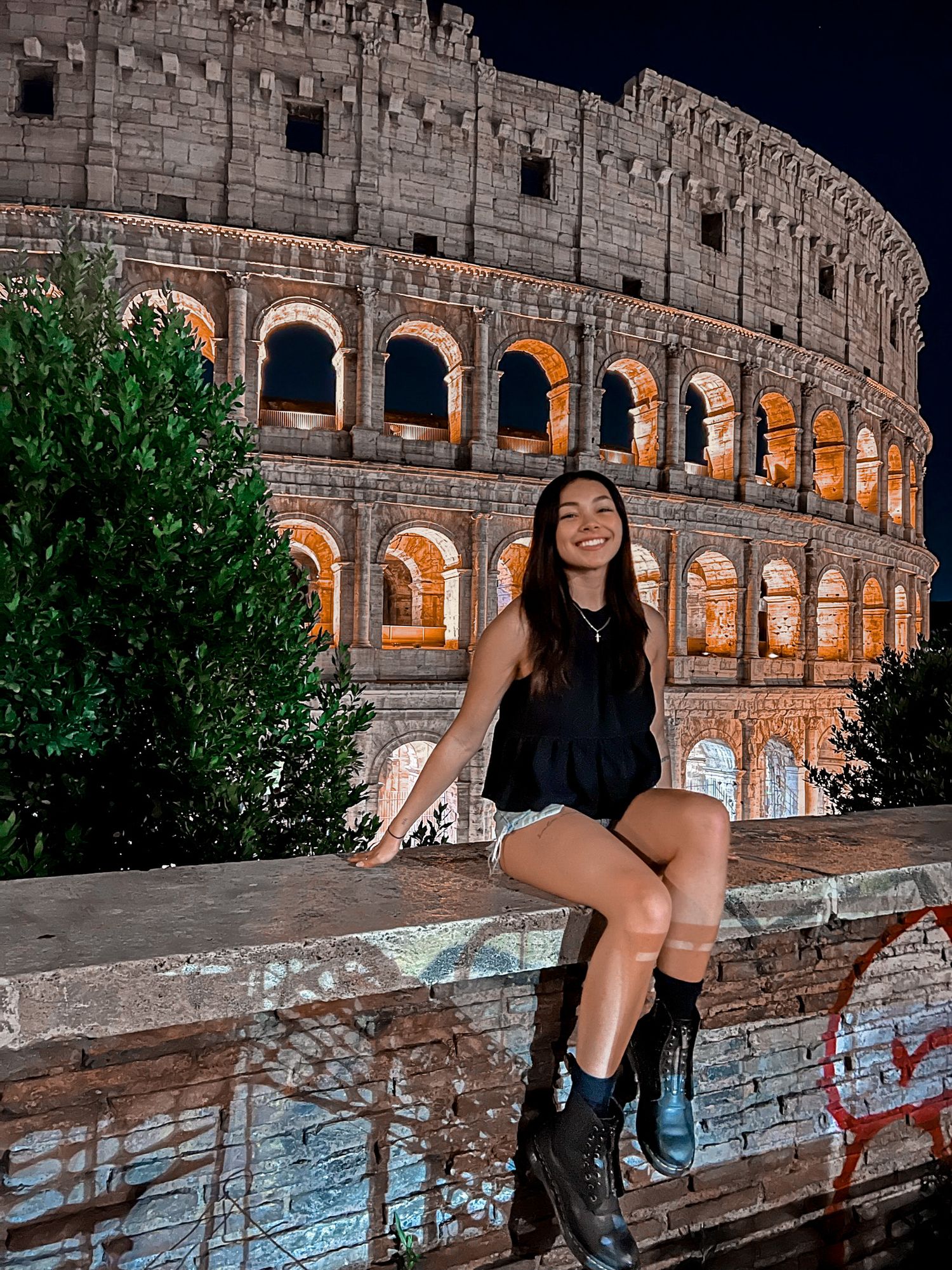 Smiling woman sitting on a stone wall in front of the illuminated Colosseum at night.
