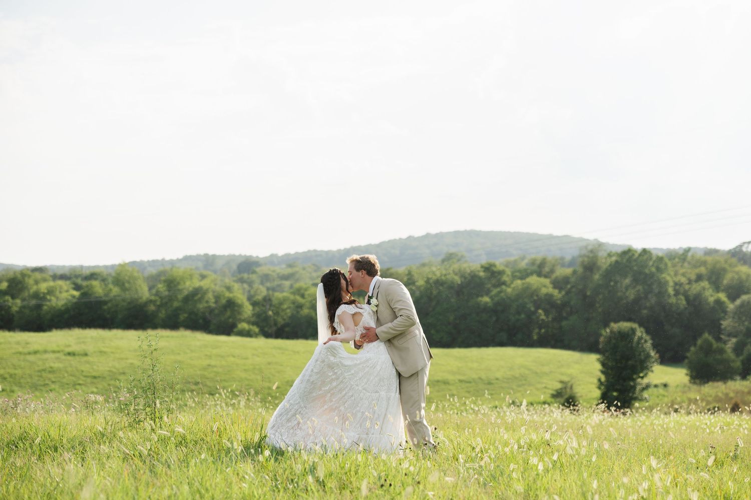A bride and groom share a romantic kiss in a lush green meadow with rolling hills in the background.