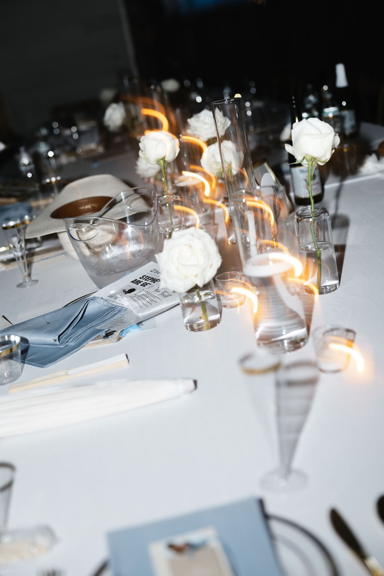 Elegant wedding table setting with white roses in glass vases, candles, and blue napkins on a white tablecloth.