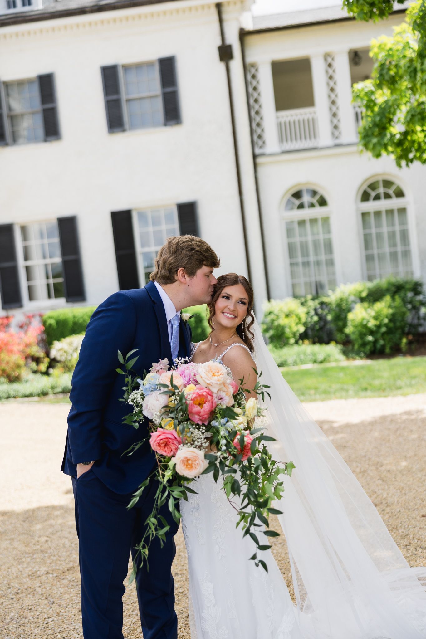 Groom in navy suit kisses bride holding lush pink floral bouquet outside elegant white manor house.