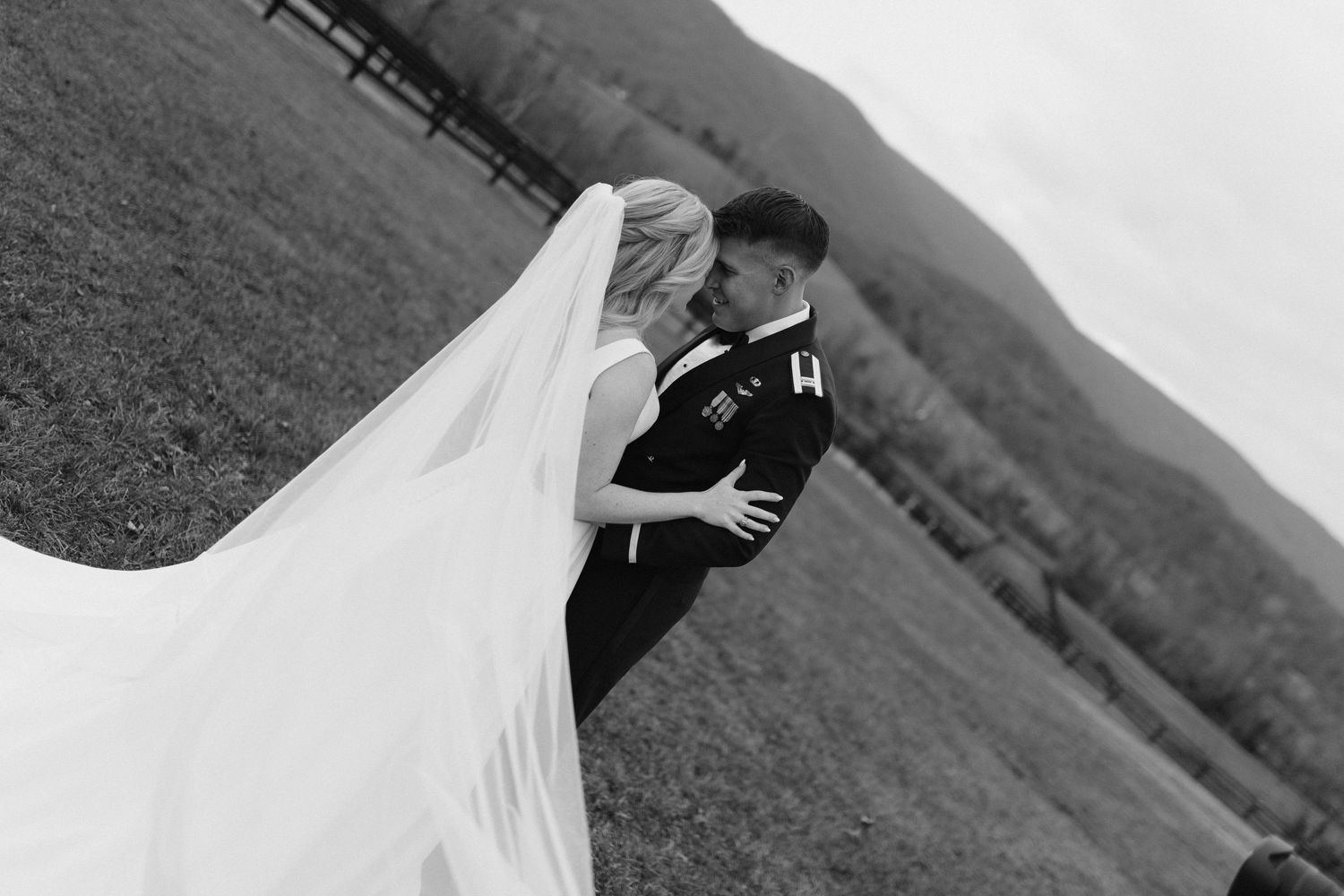 Black and white photo of bride and groom sharing romantic kiss with flowing veil in open field.