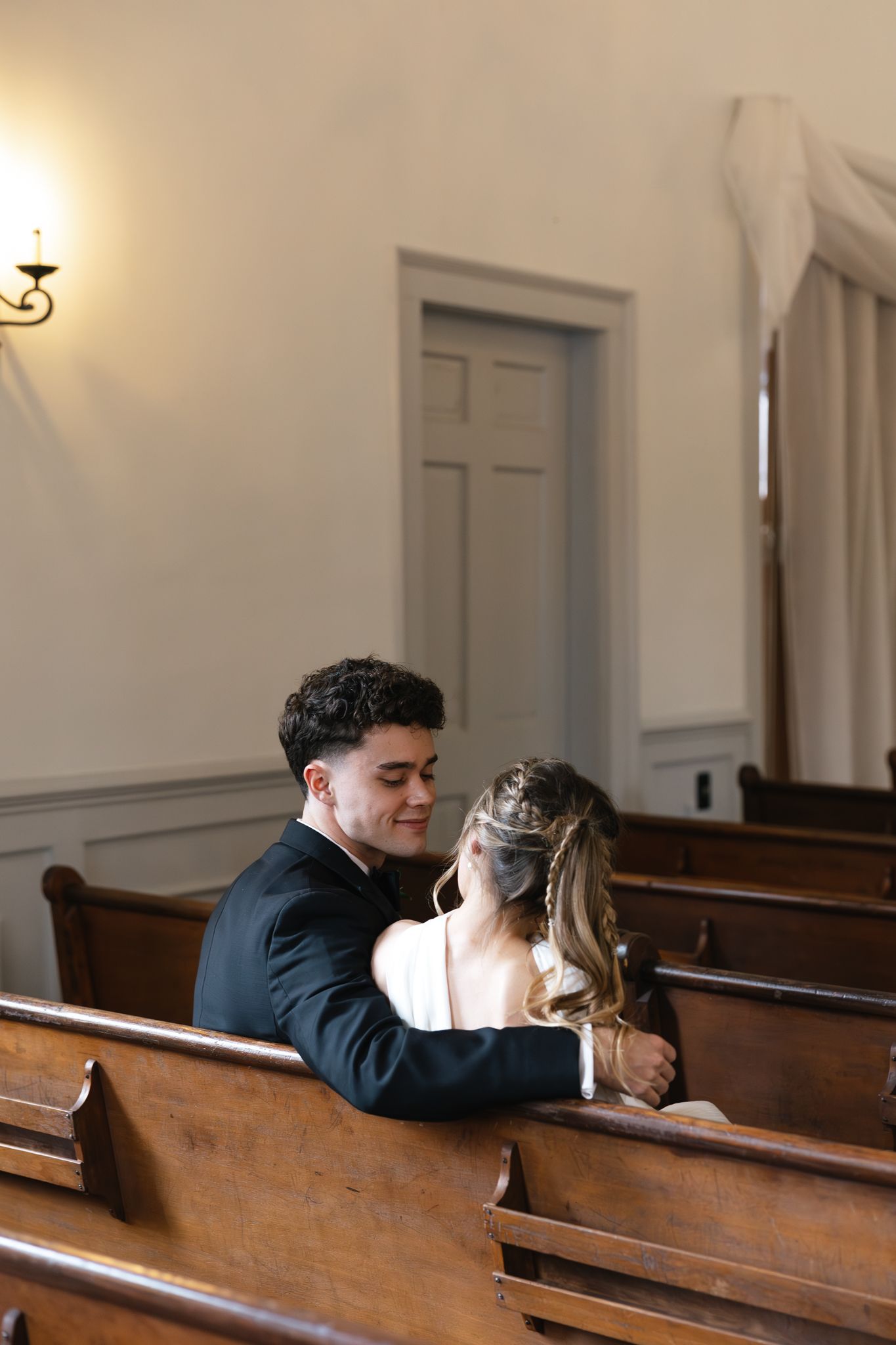 Groom in black suit embraces smiling bride in white gown while seated in wooden church pew.