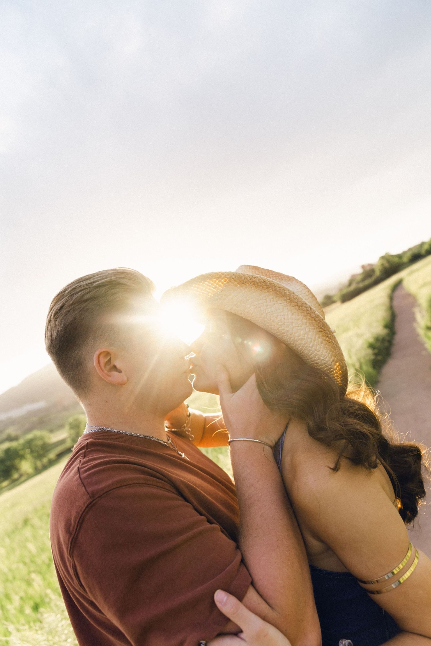 Couple sharing a sunset kiss on a scenic hillside with golden sunlight flare between them.