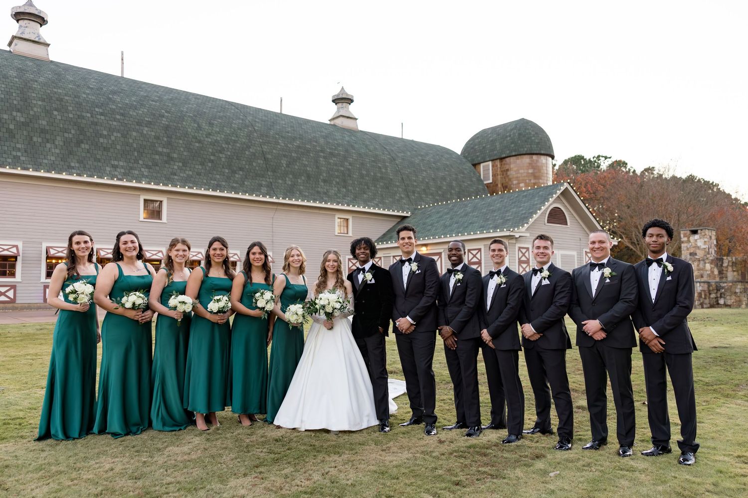 Wedding party posing in emerald green bridesmaid dresses and black tuxedos outside rustic barn venue.