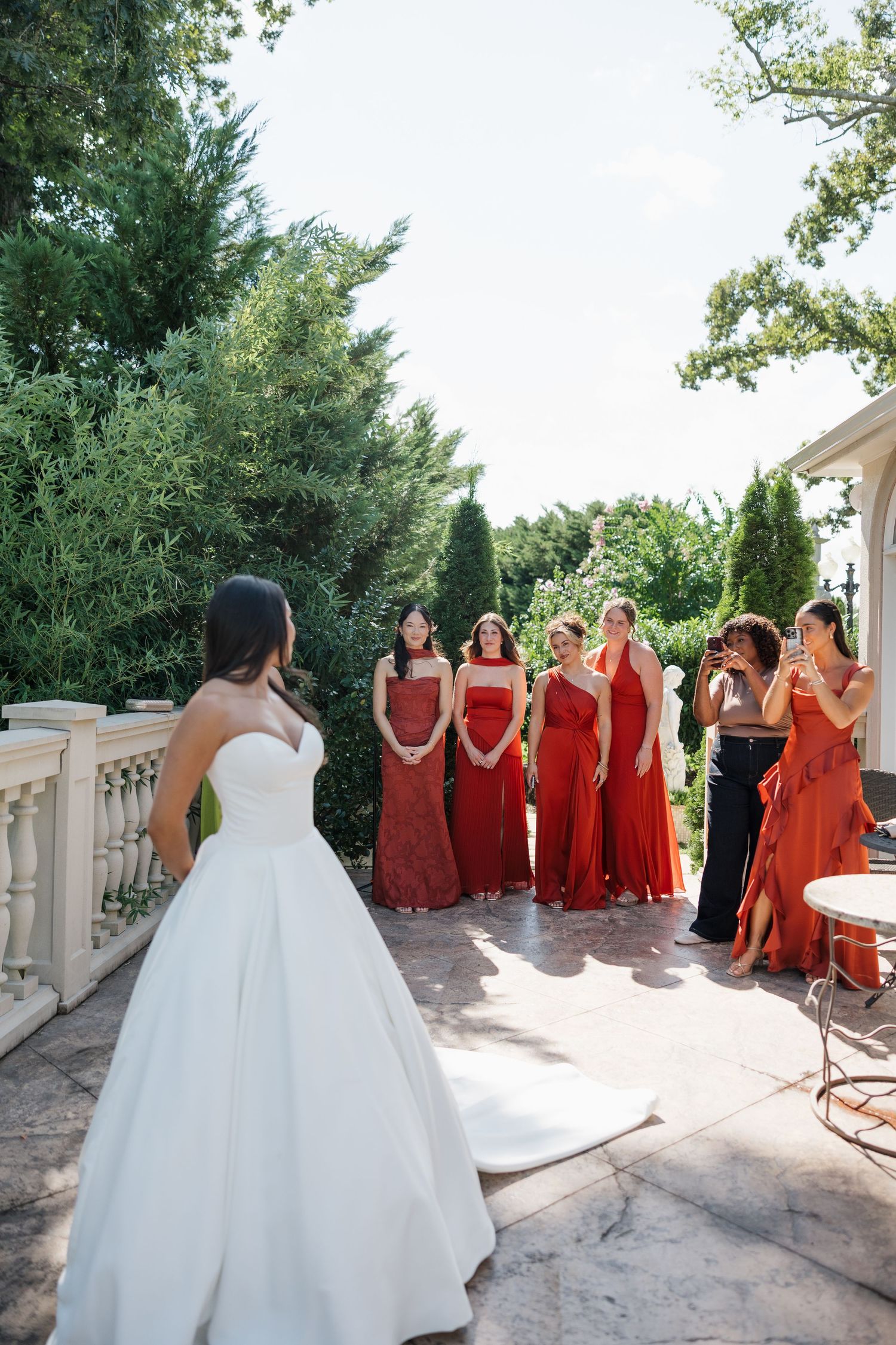 Bride in white gown reveals look to bridesmaids in red dresses during outdoor first look at garden wedding.