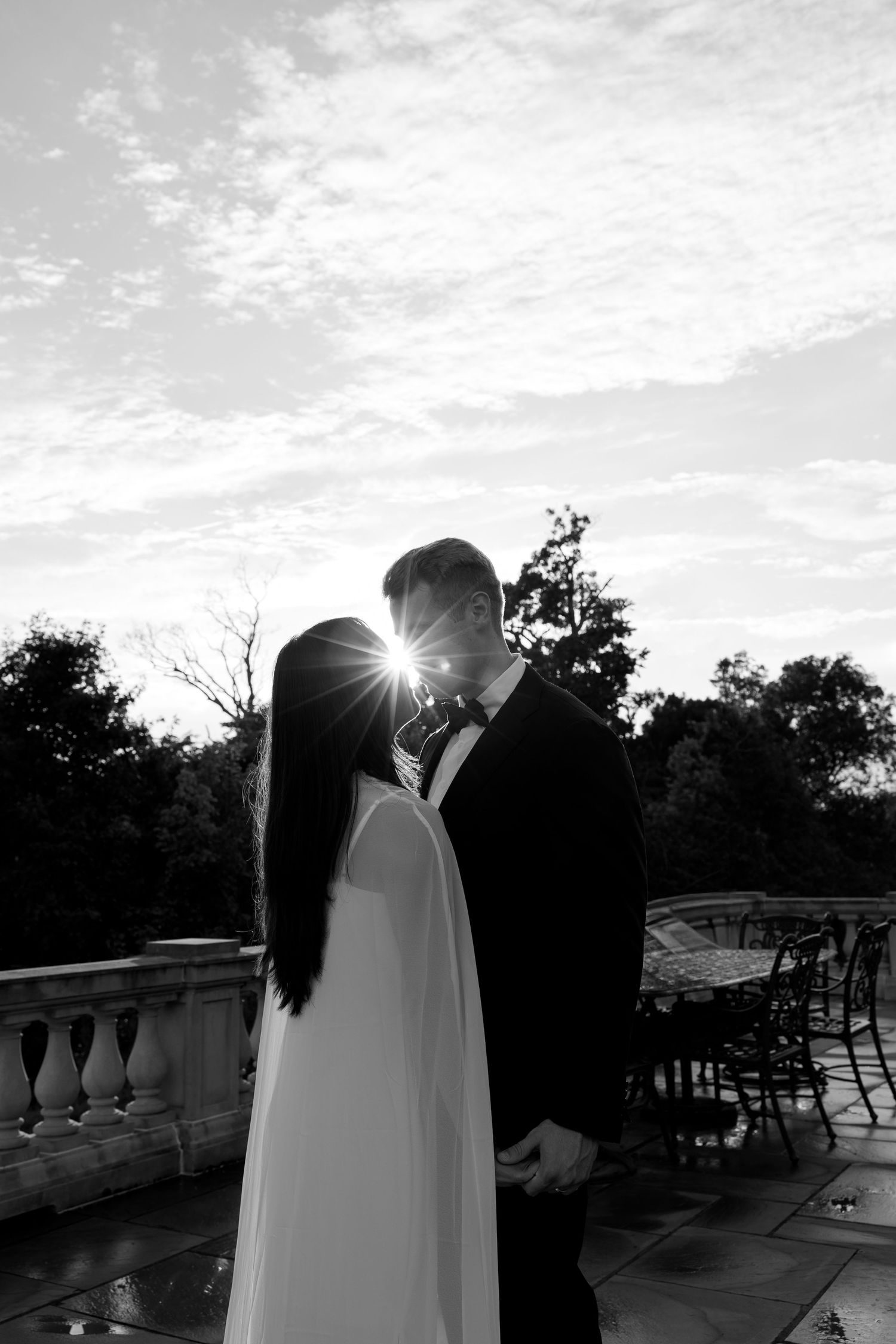 Black and white photo of a bride and groom sharing a romantic moment on a terrace, with sunlight glowing between them.