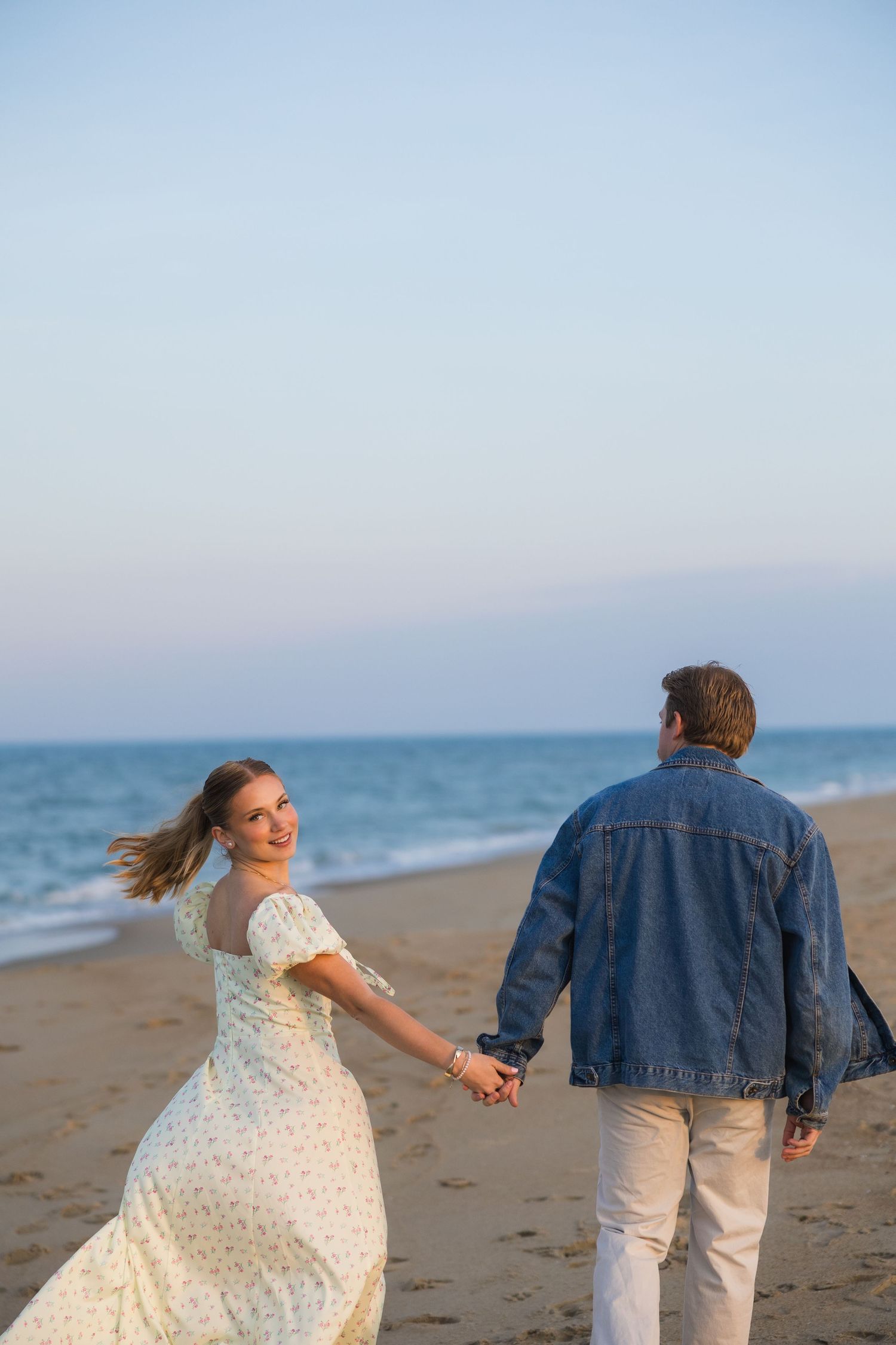 Woman in floral dress looks back smiling while holding hands with man in denim jacket walking along beach at sunset.