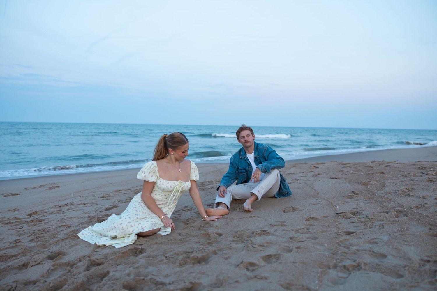Couple kneeling together on sandy beach at dusk, facing each other near ocean waves.