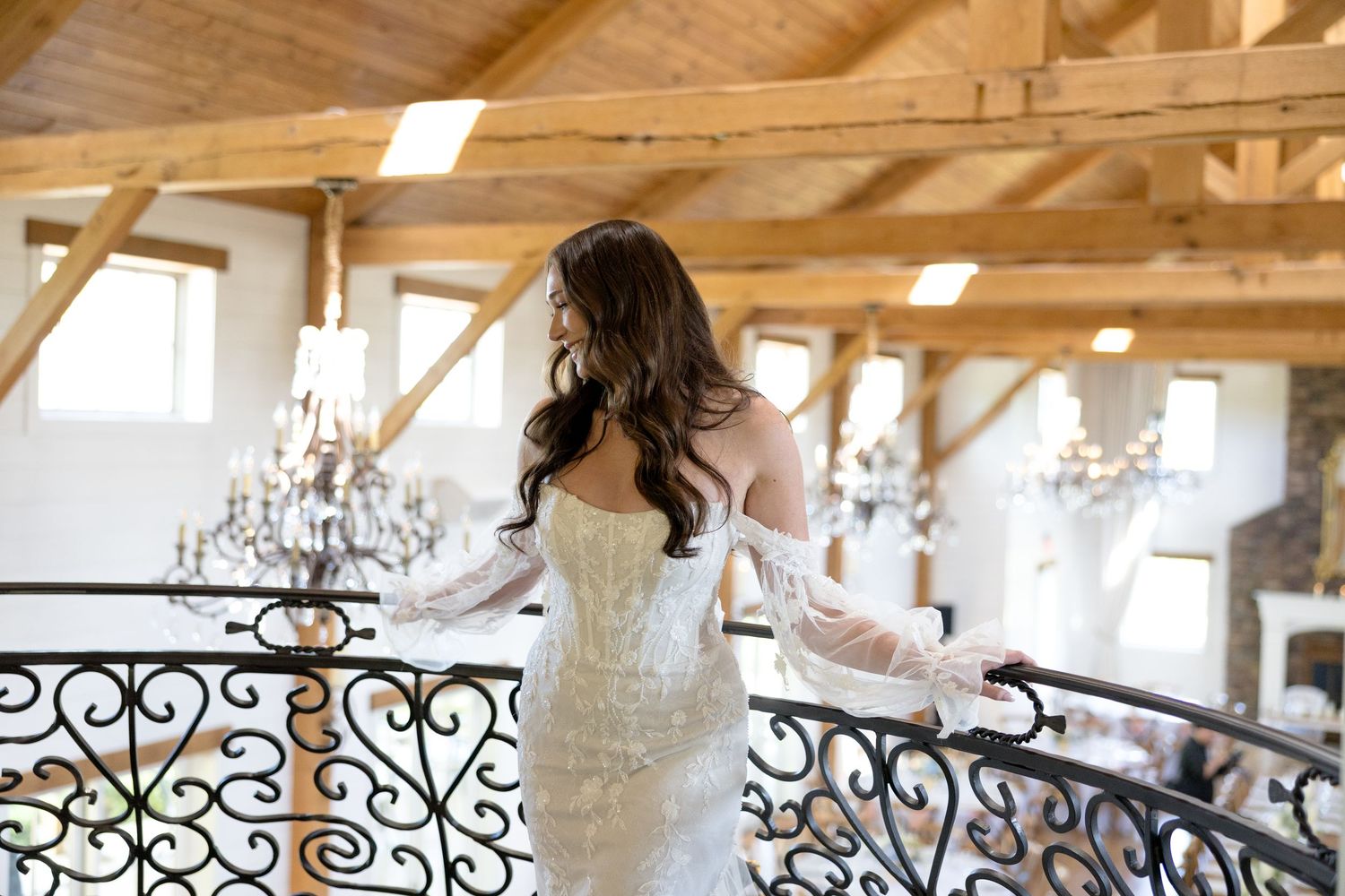Bride in fitted lace gown standing on balcony in rustic barn venue.