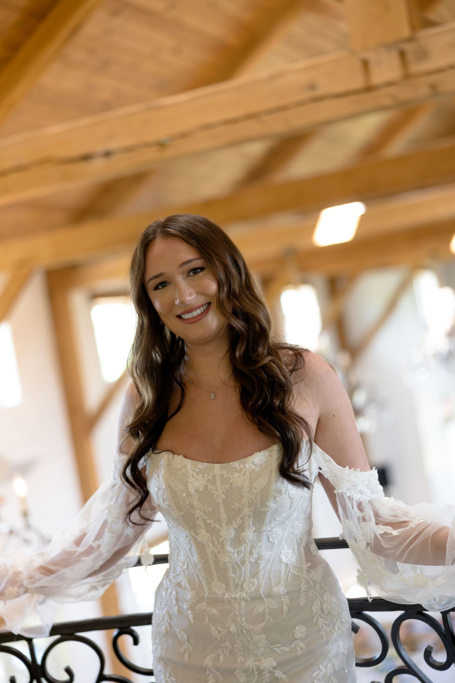 Smiling bride in a strapless lace wedding gown with sheer sleeves poses on a balcony under wooden beam ceiling.