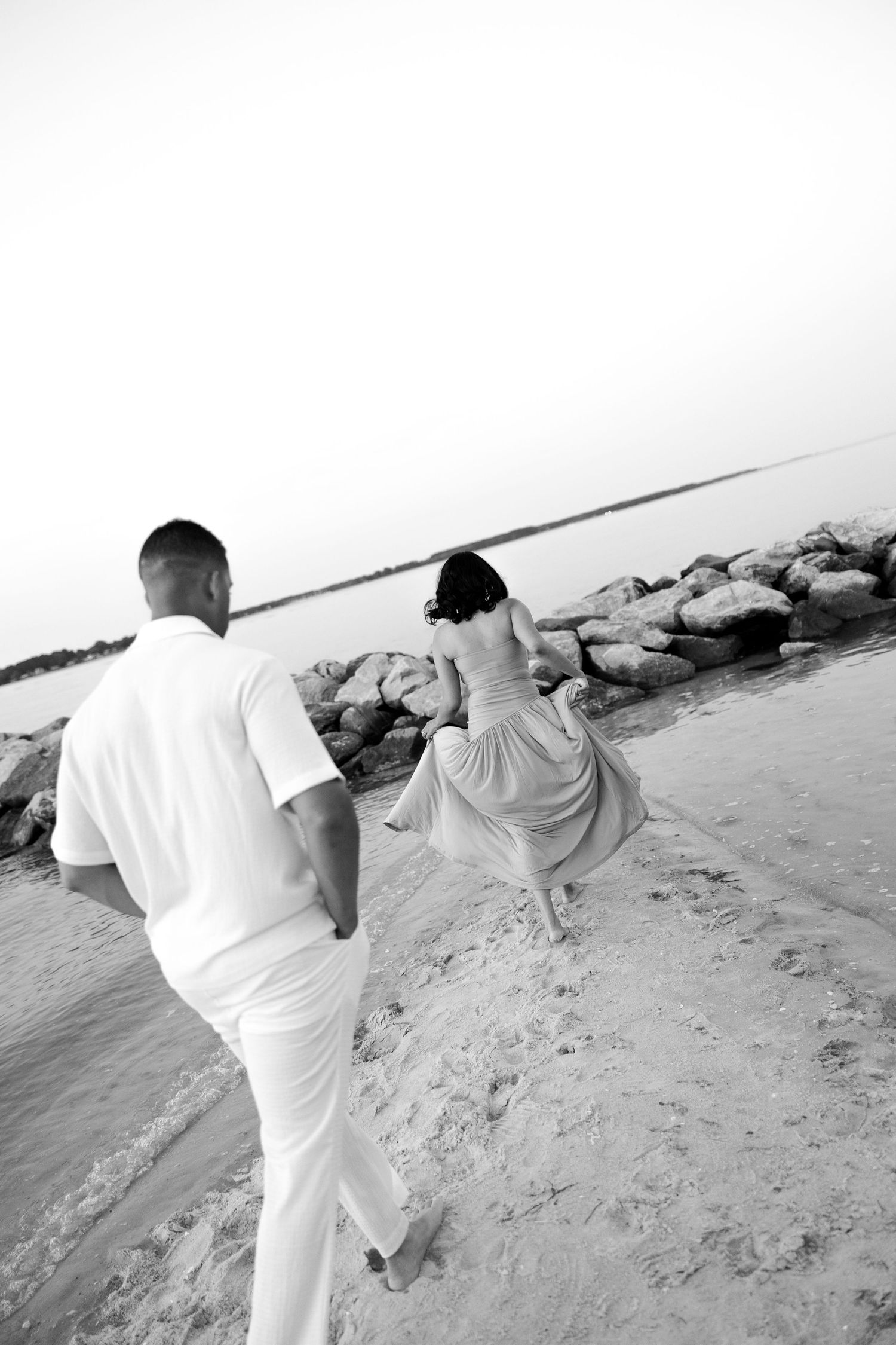 Black and white photo of a couple walking barefoot on a sandy beach toward rocky shoreline at sunset.