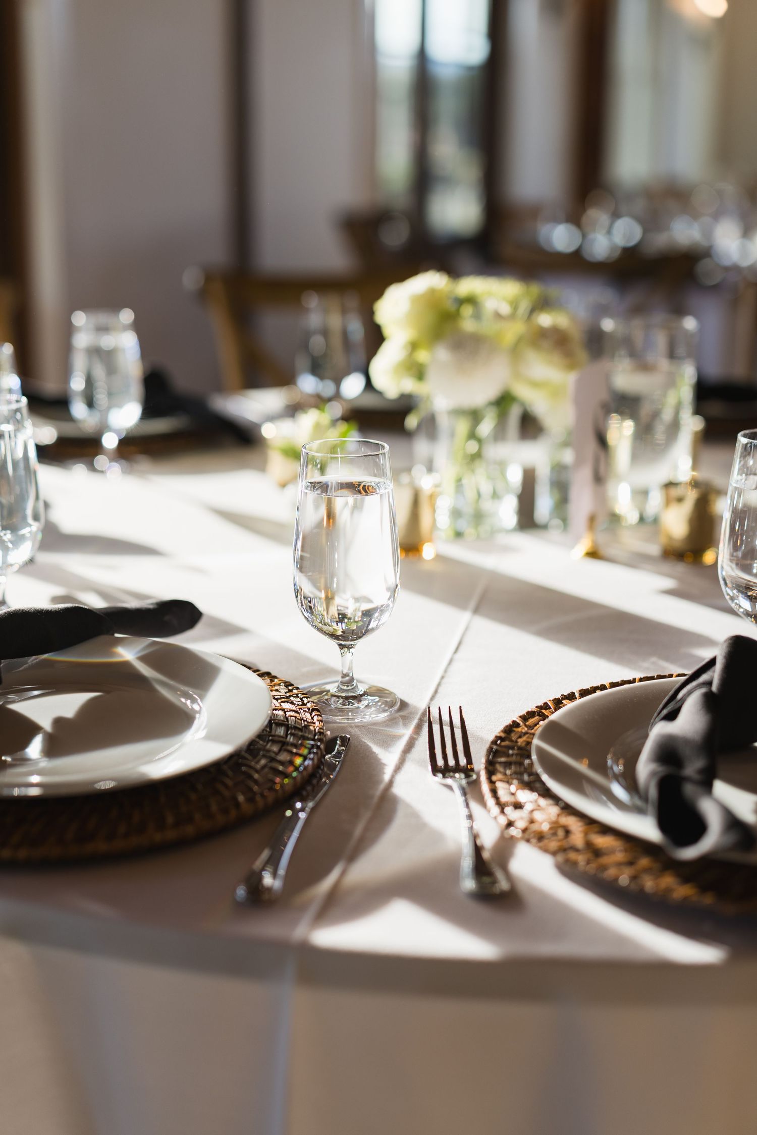 Sunlit wedding reception table set with ornate chargers, black napkins, crystal glasses, and floral centerpiece.