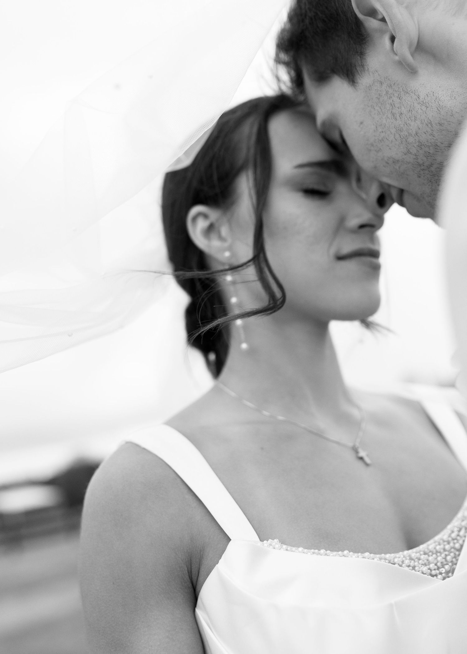 Black and white close-up of bride with veil flowing as groom tenderly kisses her forehead.