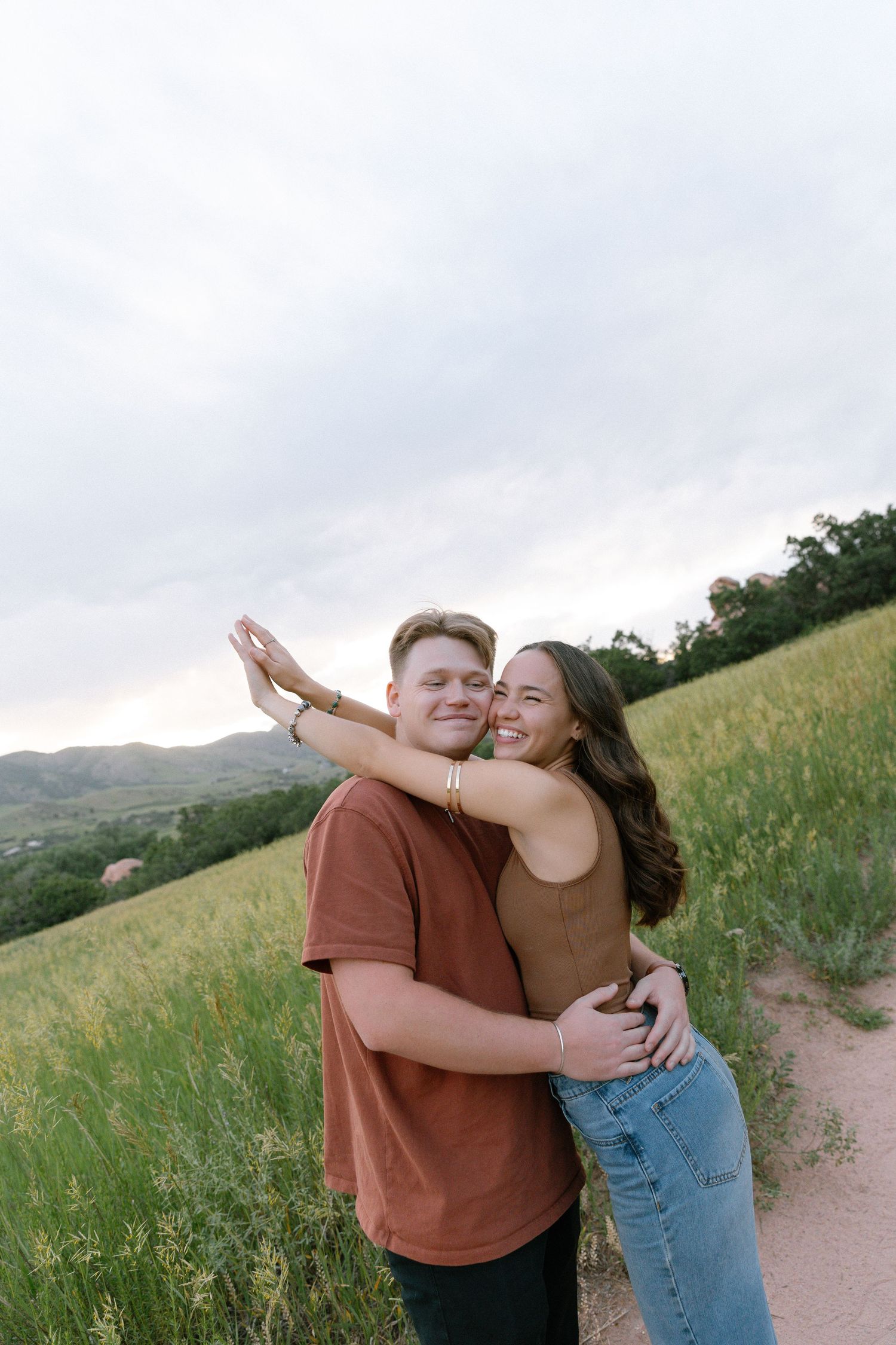 Smiling couple embracing on a dirt path surrounded by green fields and cloudy sky.