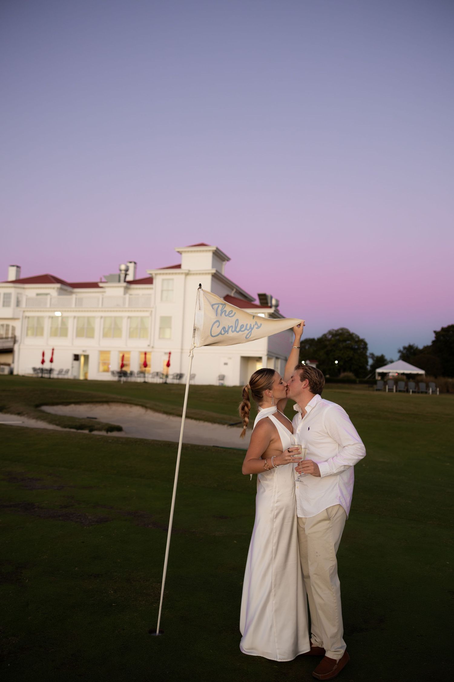 Newlywed couple kissing on a golf course at sunset, holding a wedding flag, with a white venue building behind them.