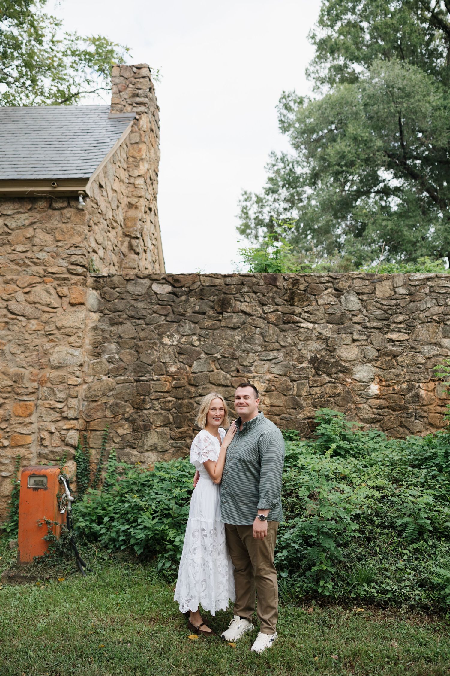 A couple poses together in front of a rustic stone wall surrounded by lush greenery, woman in white lace dress.