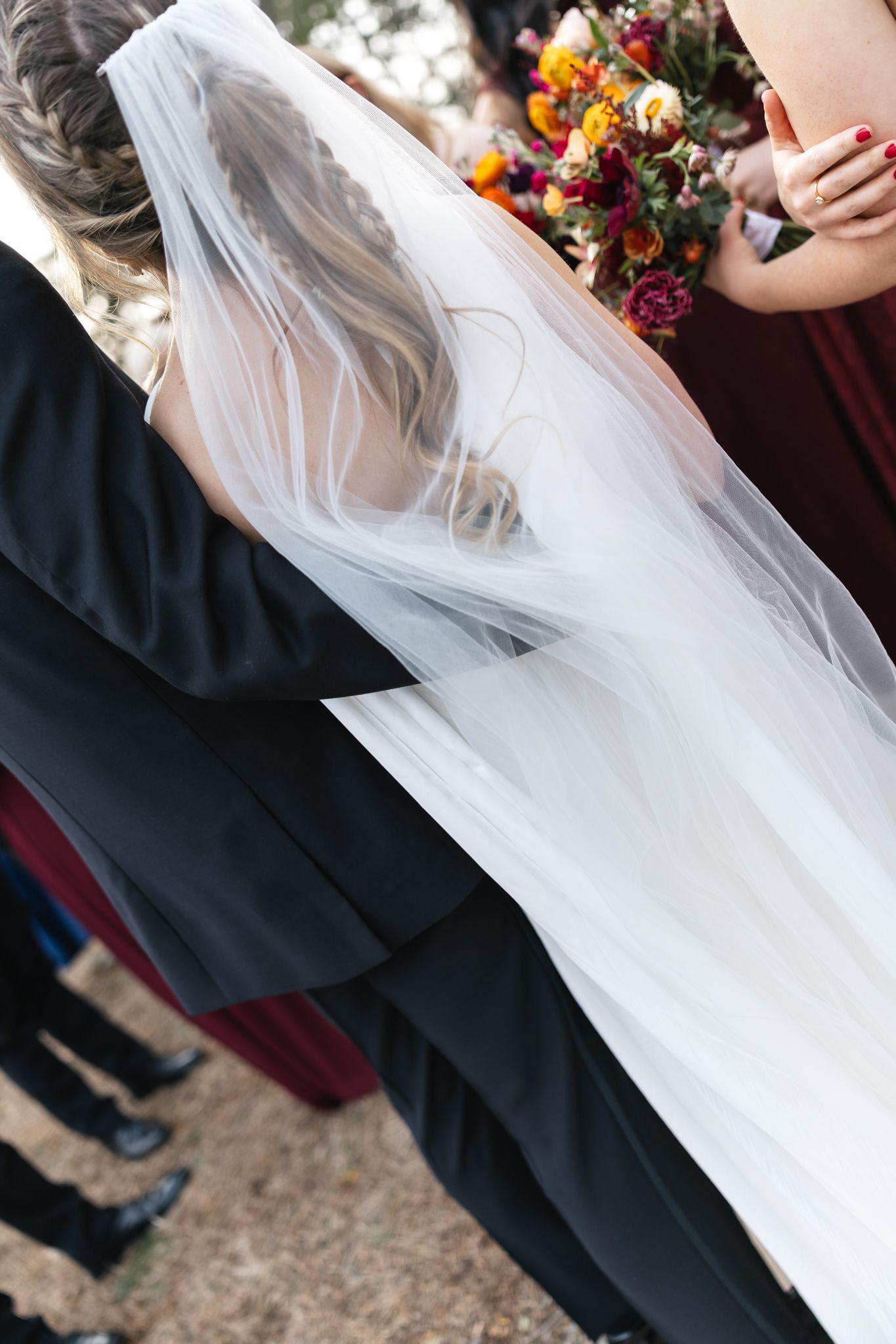 Close-up of bride's flowing veil draped over groom's black tuxedo during outdoor wedding ceremony.