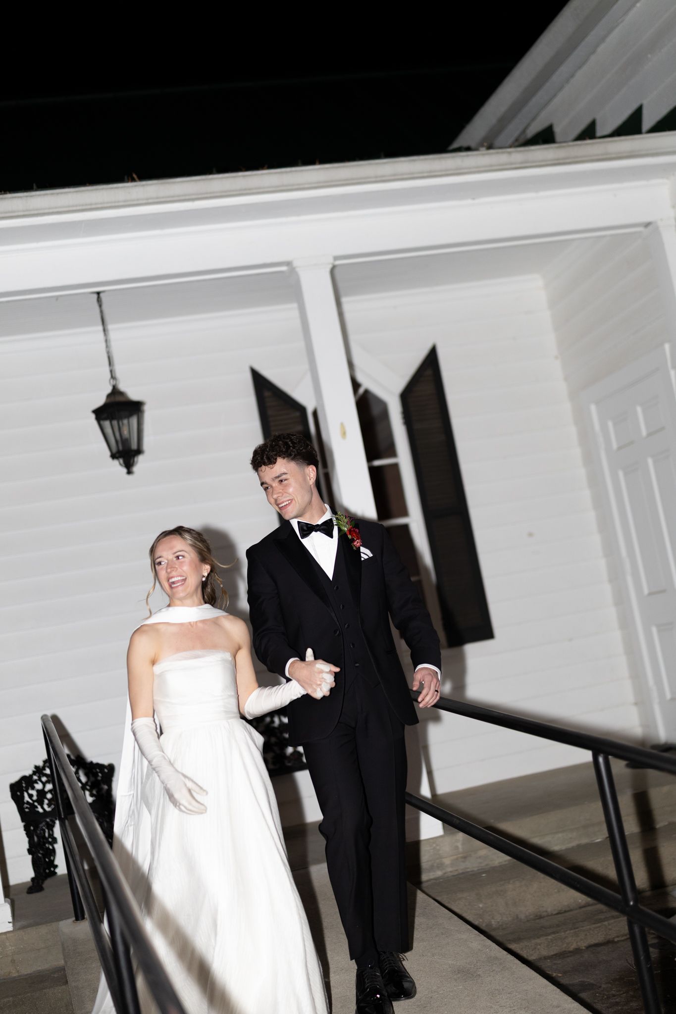 Bride and groom walking arm in arm outside a white chapel at night.