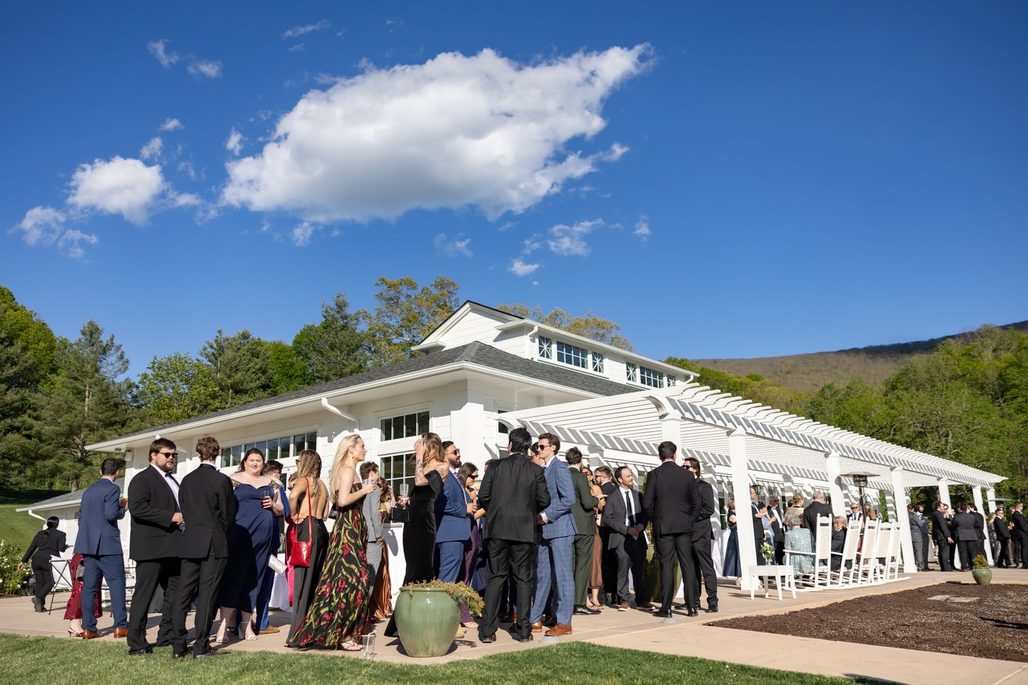 Guests mingle outdoors at a white pergola wedding venue surrounded by lush green hills under a bright blue sky.
