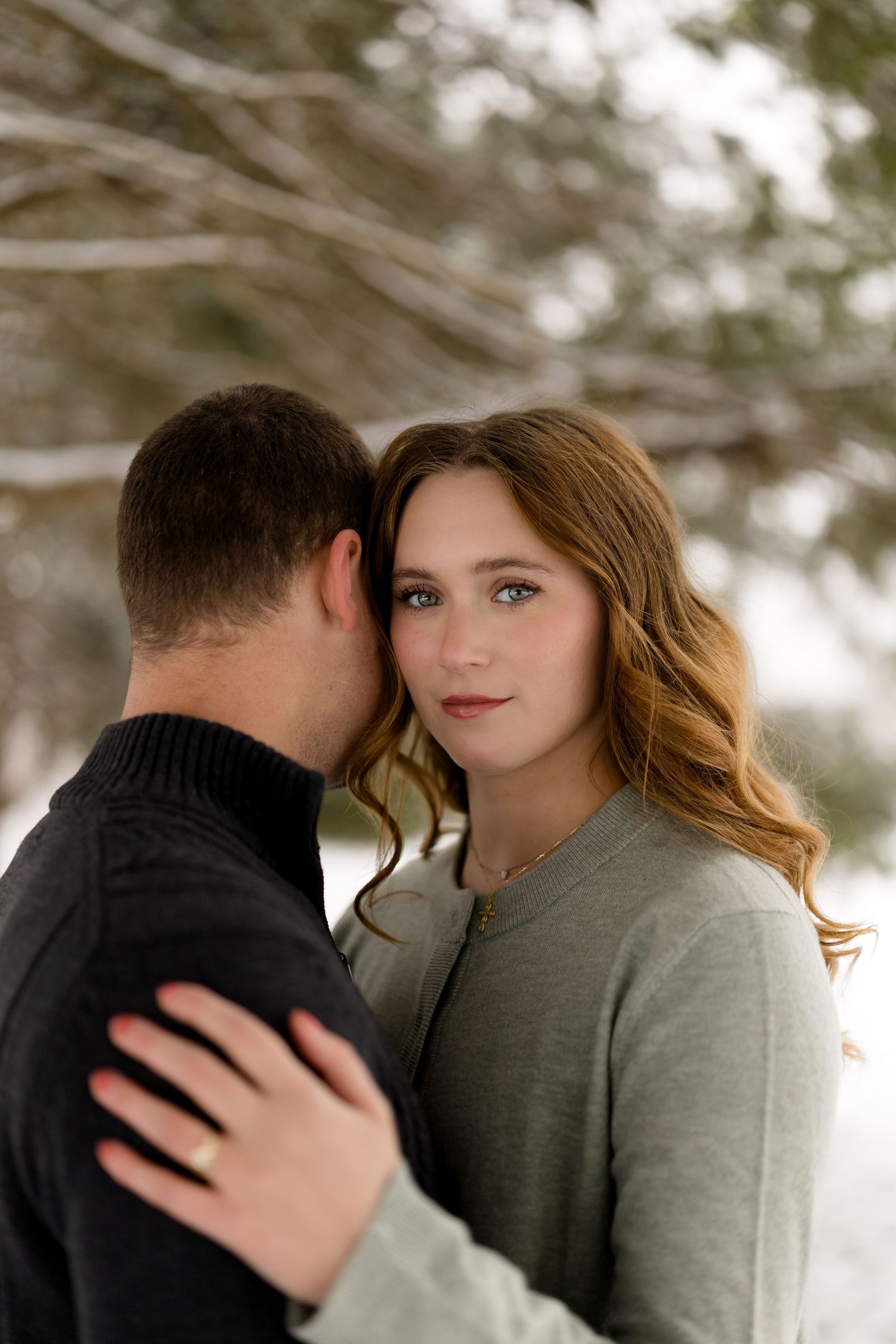 Woman with auburn hair embraces man outdoors in snowy winter landscape, gazing at camera.