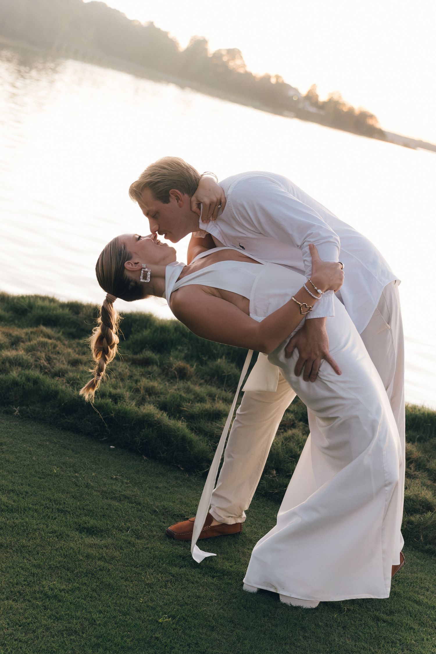 Couple in white outfits sharing a romantic dip kiss by a sunlit waterfront on their wedding day.
