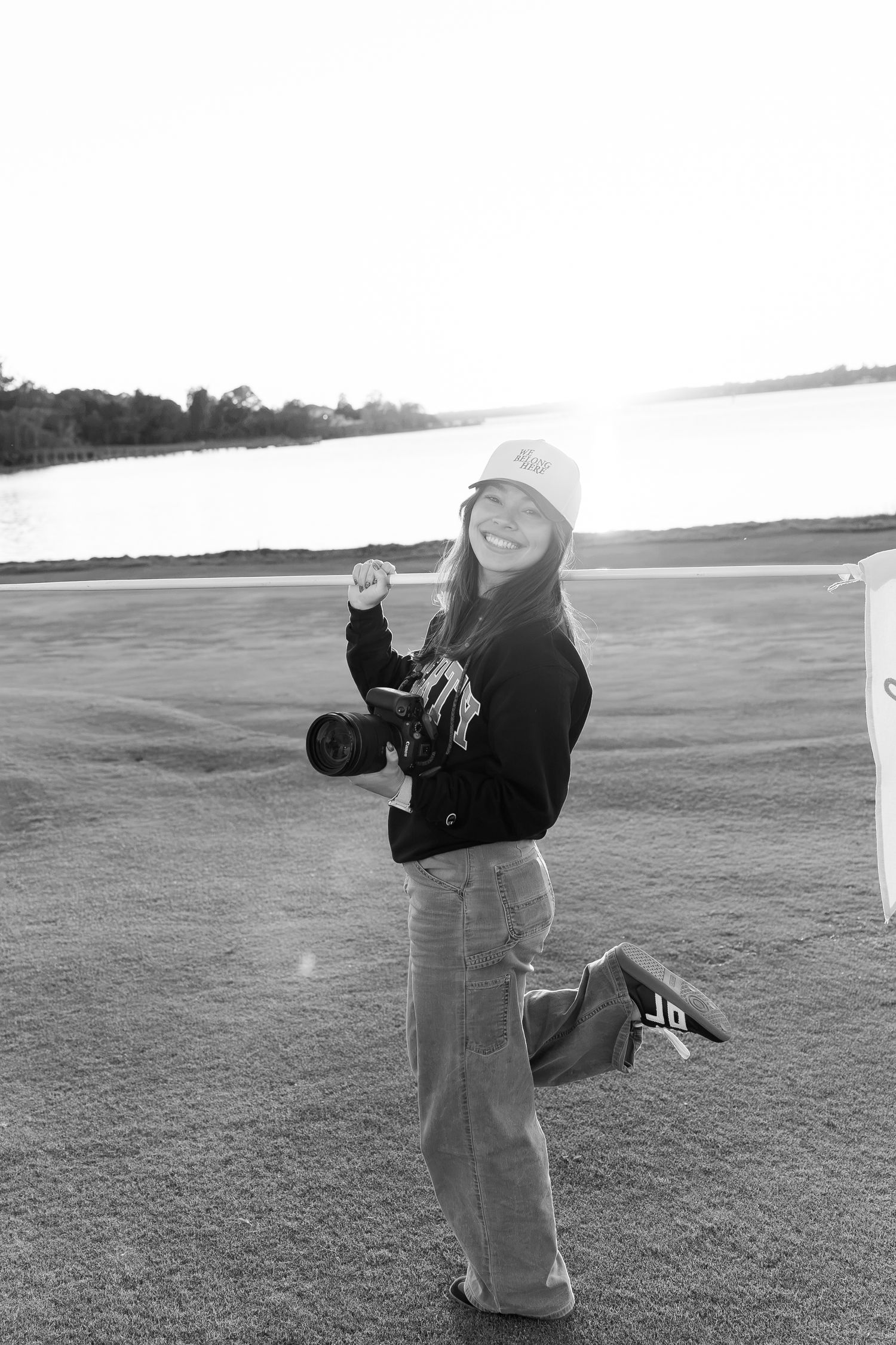 Black and white photo of a smiling woman holding a camera outdoors near a lake, wearing a hoodie and wide-leg jeans.