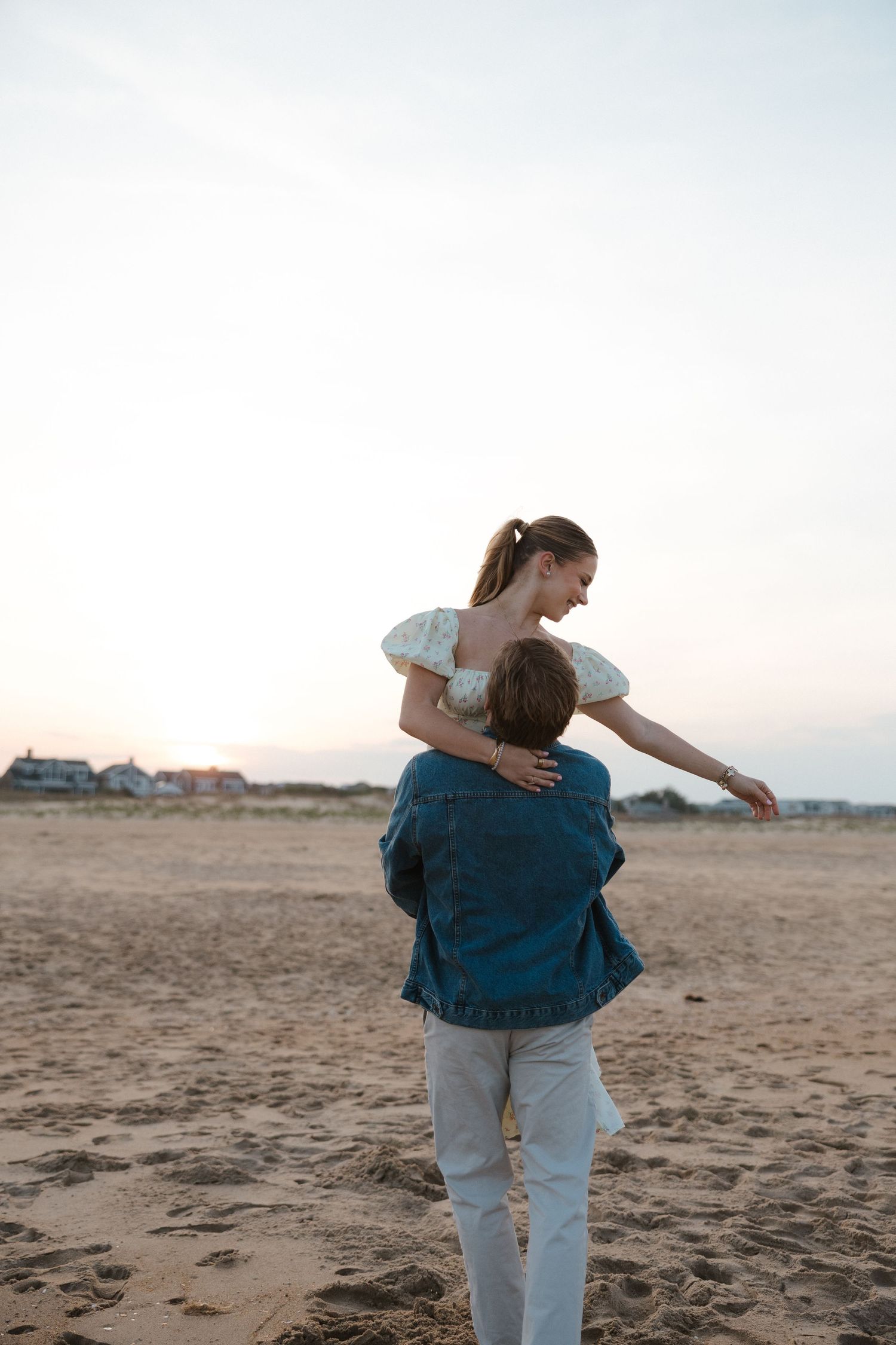 Man in denim jacket lifts woman in white floral dress on sandy beach at dusk with coastal houses in background.