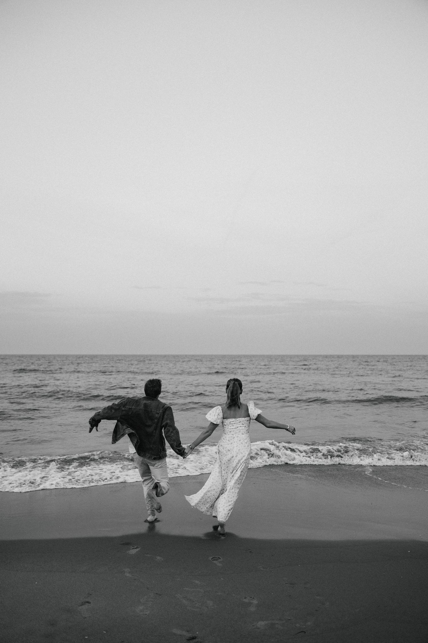 Couple holds hands running toward ocean waves on beach at sunset in black and white.