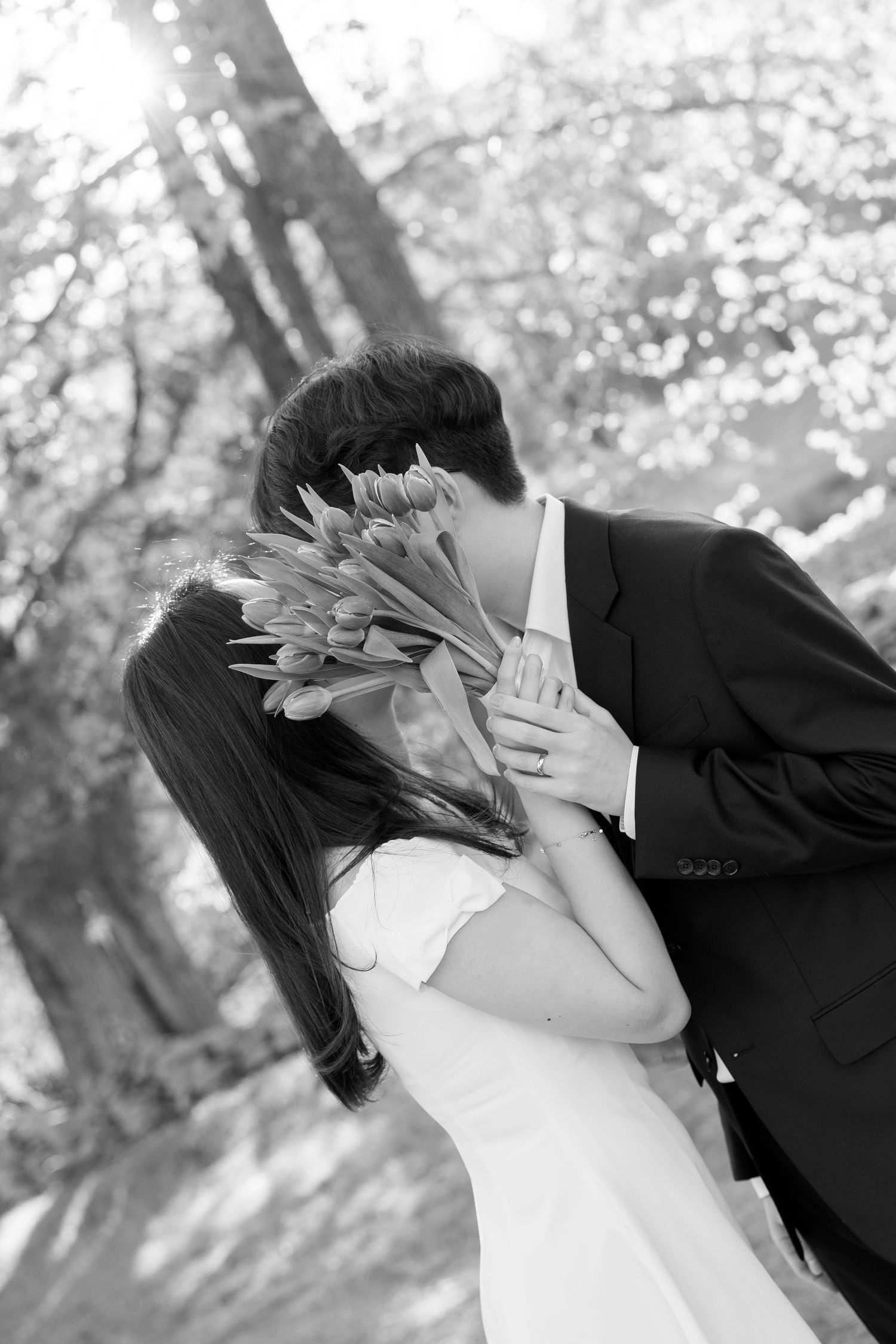 Black and white photo of a couple kissing behind a bouquet of tulips in a blooming outdoor setting.