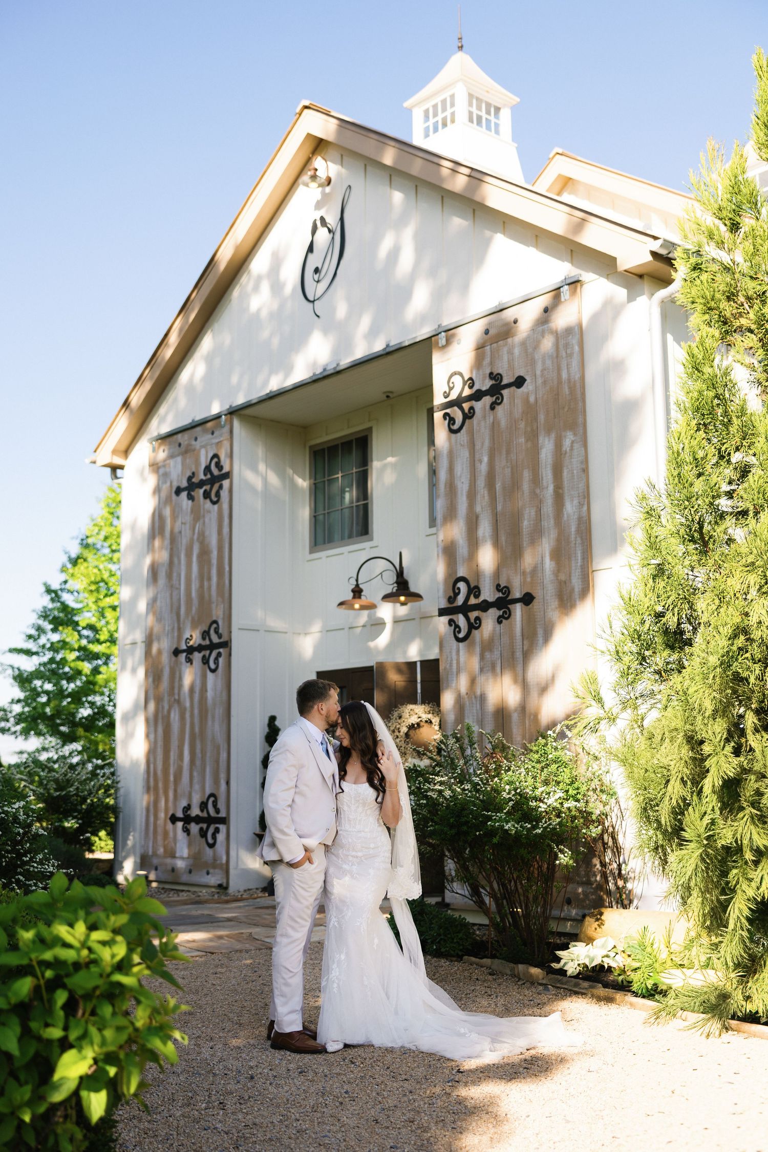 Bride and groom share a romantic kiss outside a rustic white barn wedding venue surrounded by greenery.