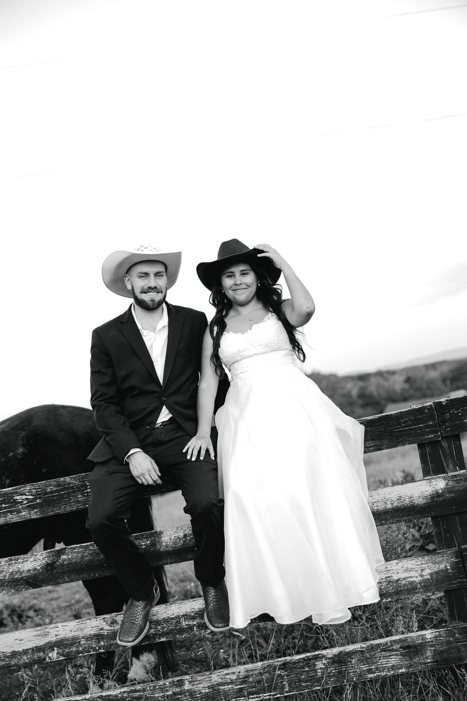 Black and white photo of a bride and groom in cowboy hats posing by a rustic wooden fence at their country wedding.