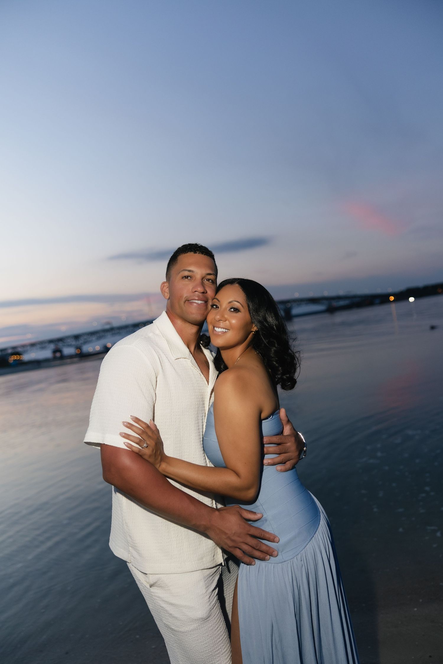 Couple embracing romantically on a beach at sunset, wearing elegant white and blue outfits with ocean backdrop.