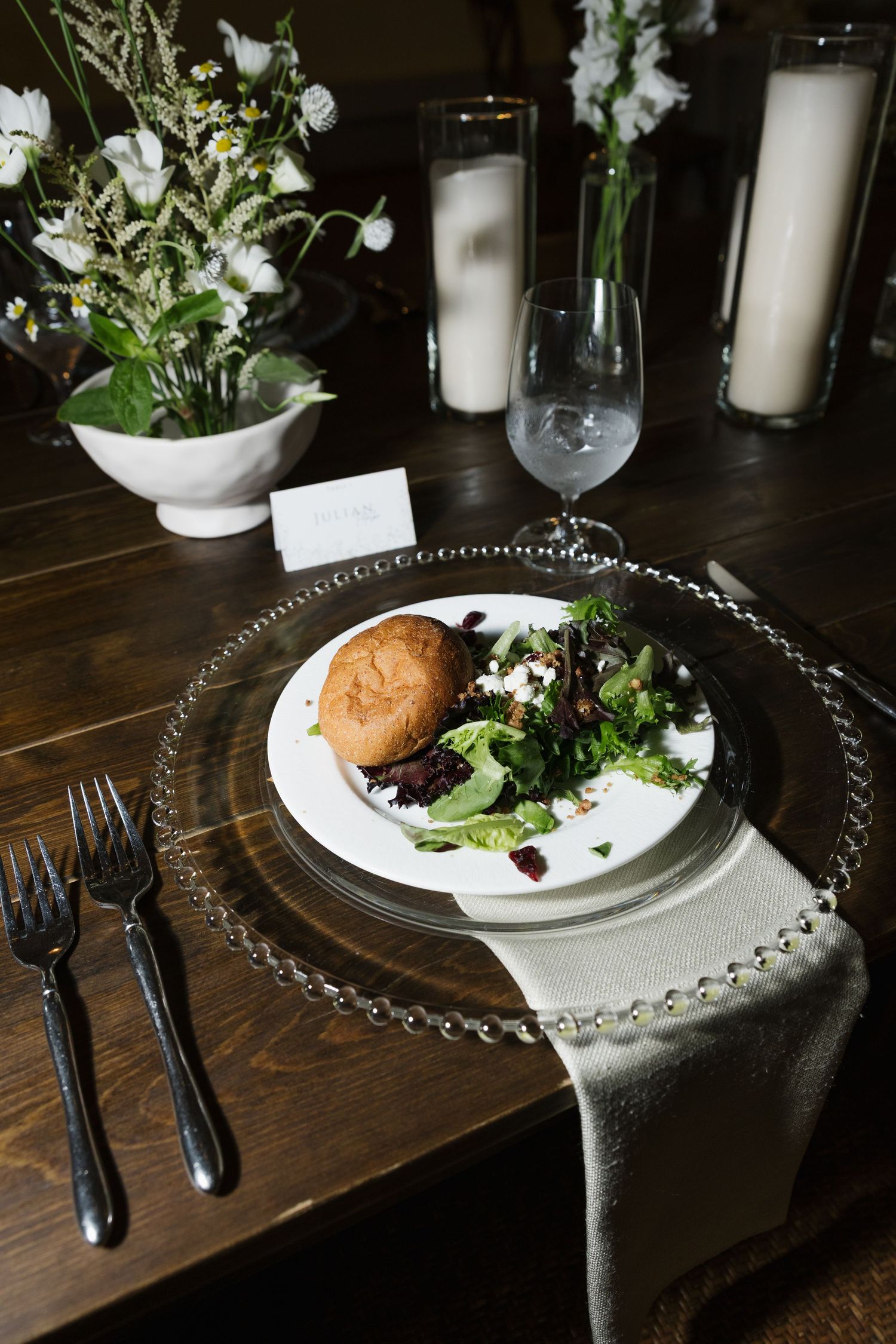 Elegant wedding table setting with salad, bread roll, glass charger plate, forks, white flowers, and candles on dark wood.