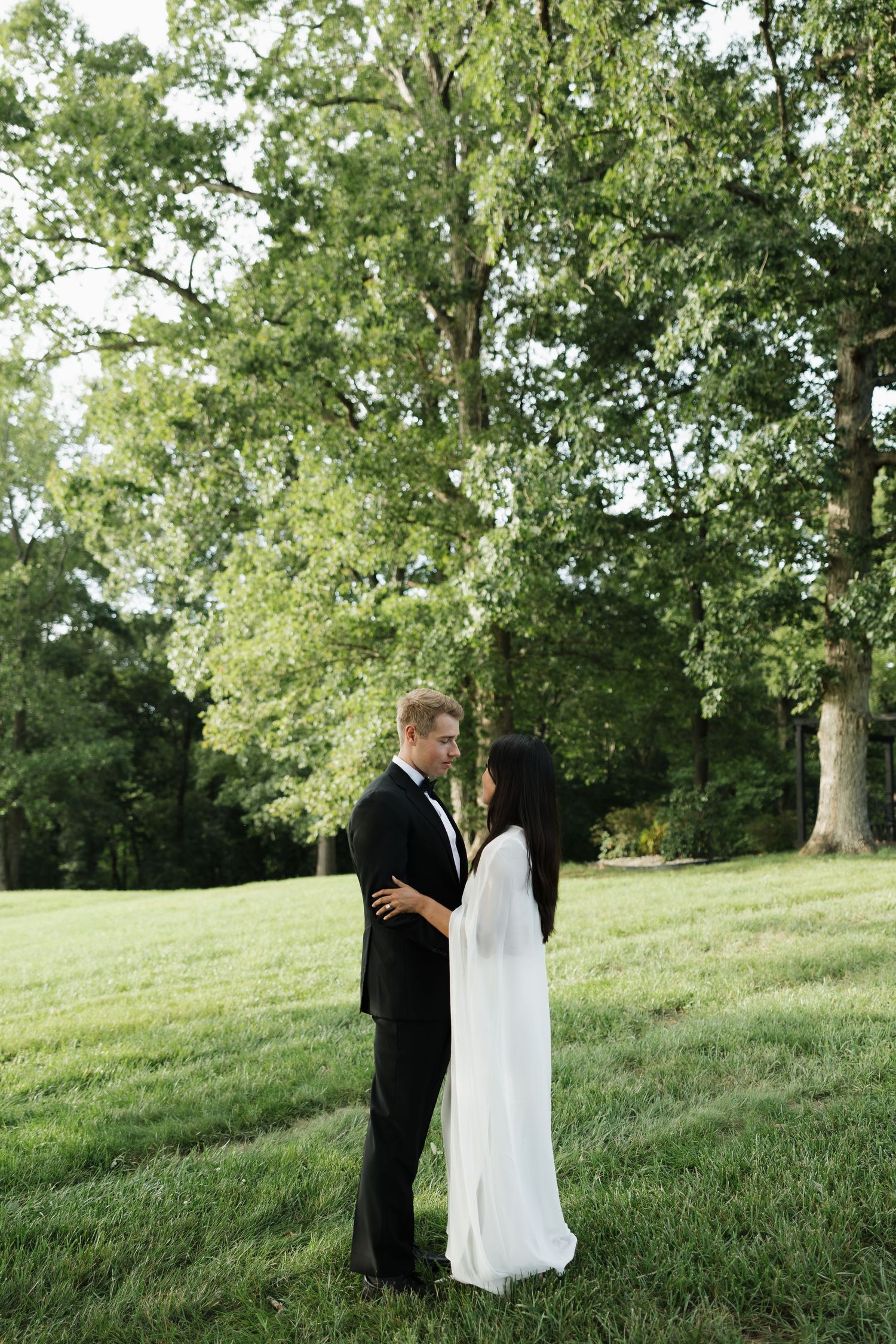 A bride and groom share a romantic moment outdoors on a lush green lawn surrounded by tall trees.