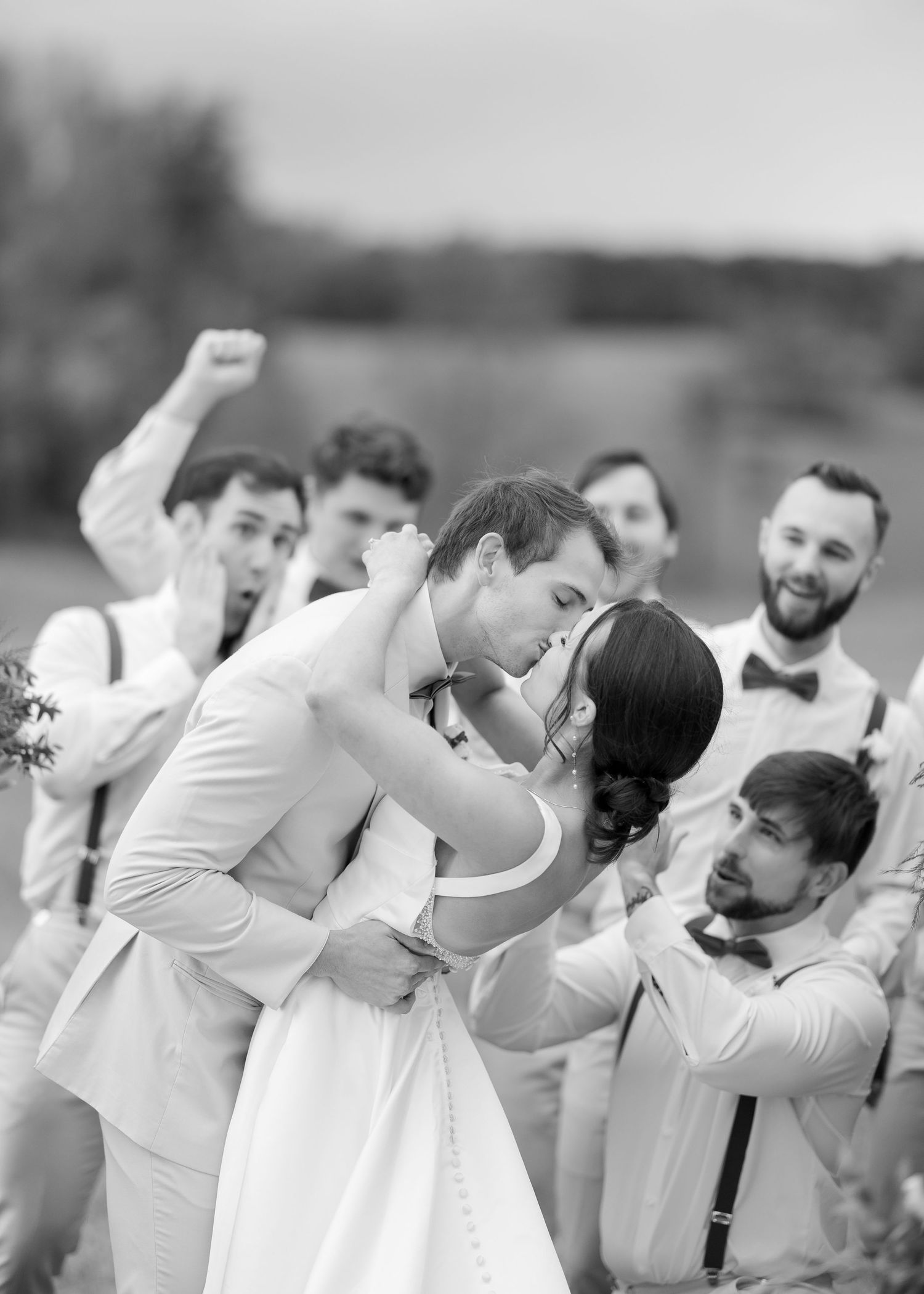 Black and white photo of groom kissing bride as excited groomsmen celebrate around them in an outdoor setting.