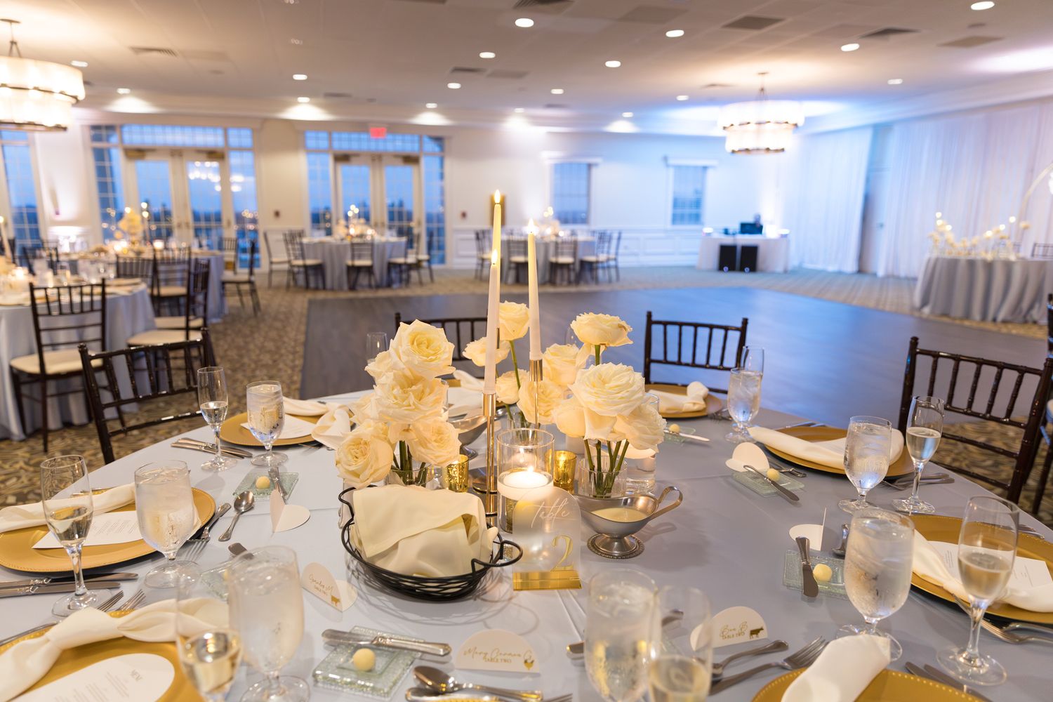 Elegant wedding reception hall with white floral centerpieces, candles, gold chargers, and chiavari chairs.