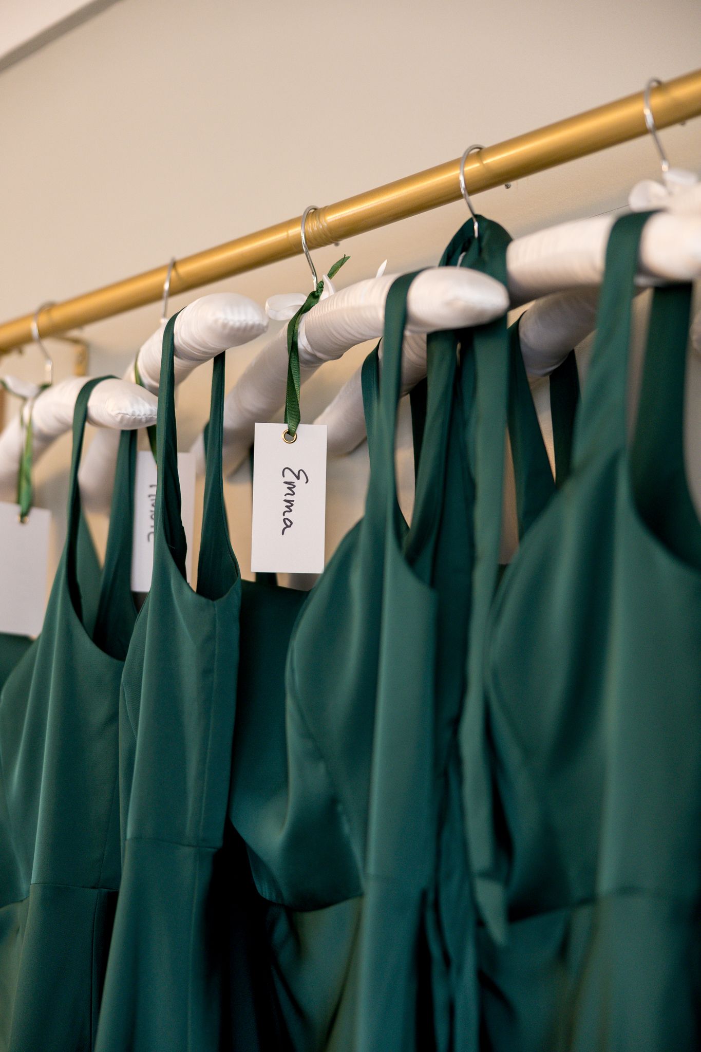 Emerald green bridesmaid dresses hanging on gold rack with white hangers and name tags in bridal suite.