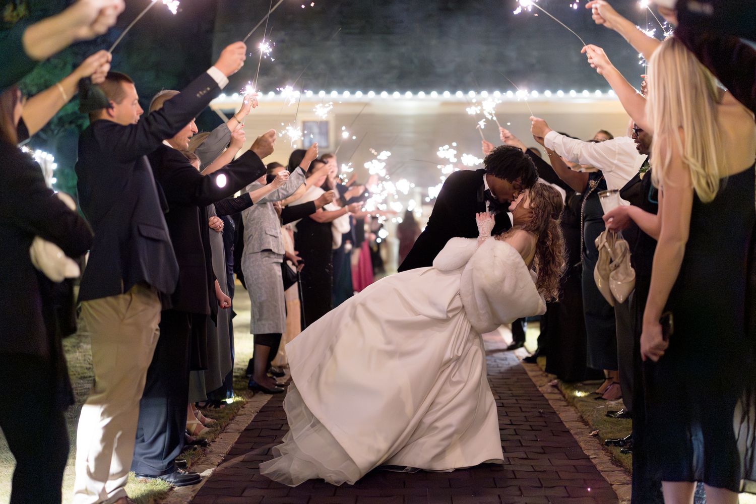 Bride and groom share a romantic dip kiss during a sparkler sendoff at their outdoor evening wedding reception.