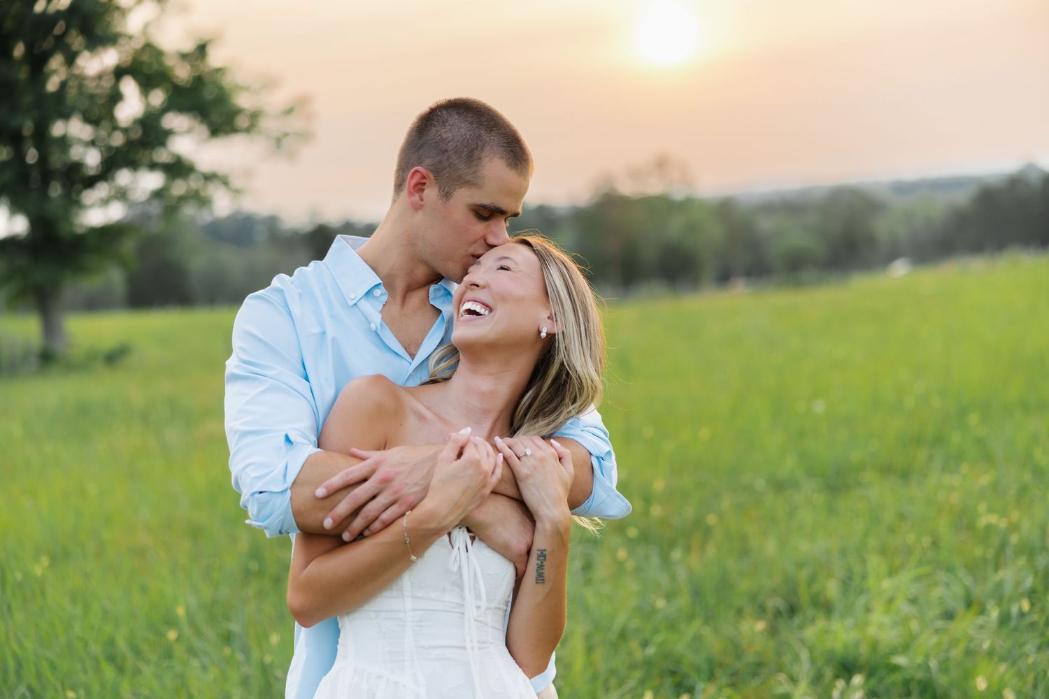 Couple embracing in a green field at sunset, smiling romantically in warm golden light.