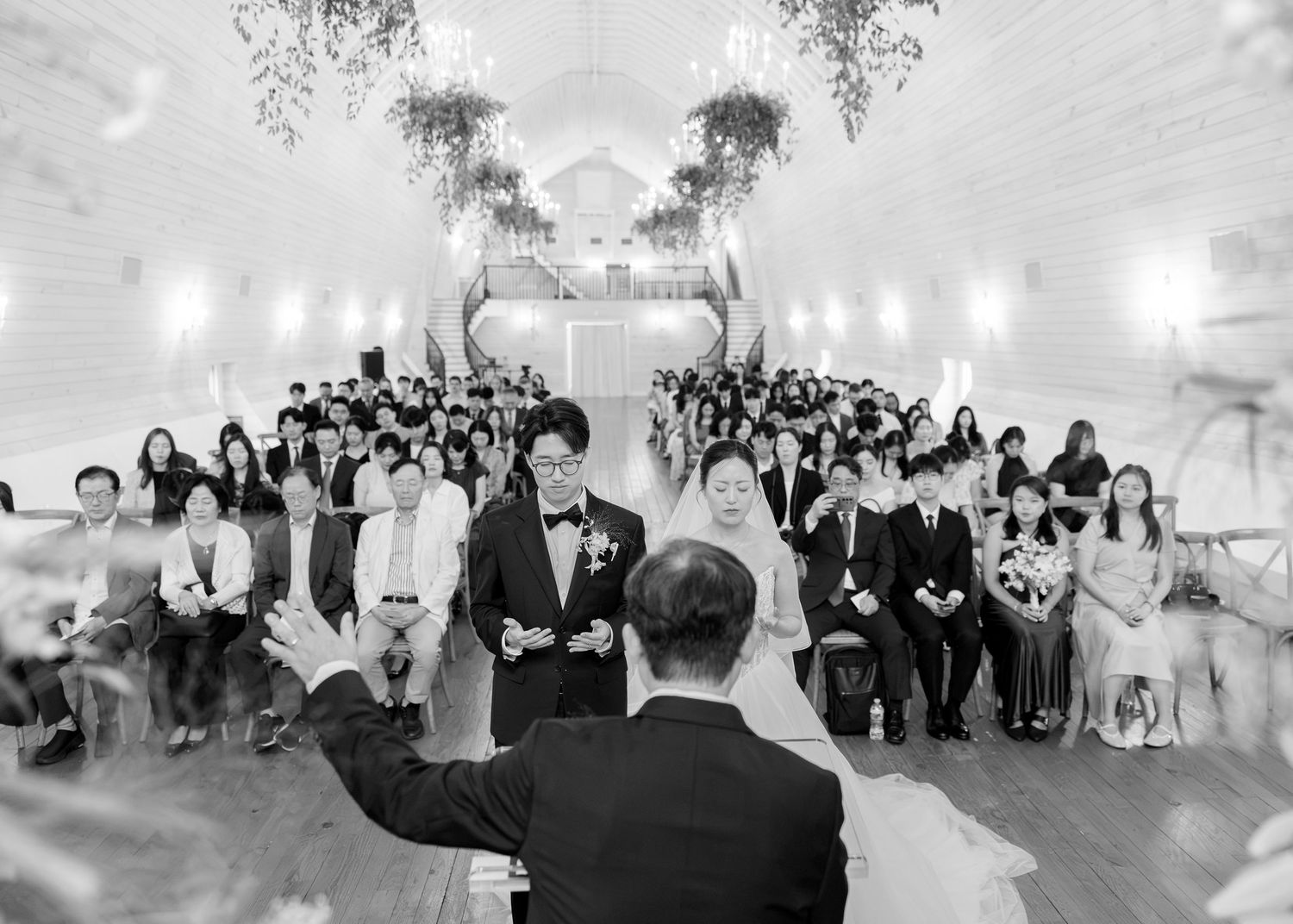 Black and white wedding ceremony photo showing bride and groom at altar with guests seated in elegant white venue.