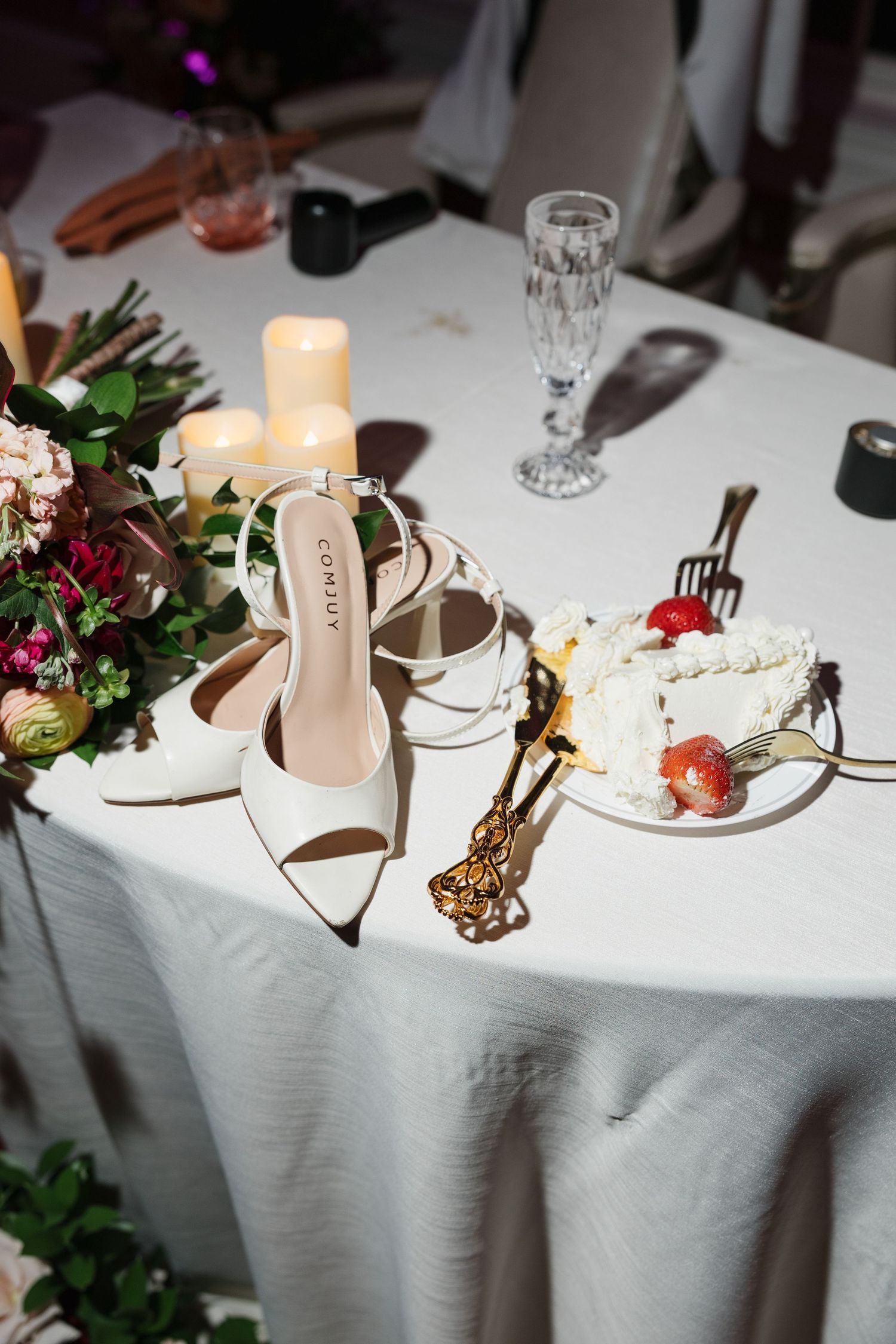 White bridal heels beside wedding cake slice, candles, and floral arrangement on reception table.