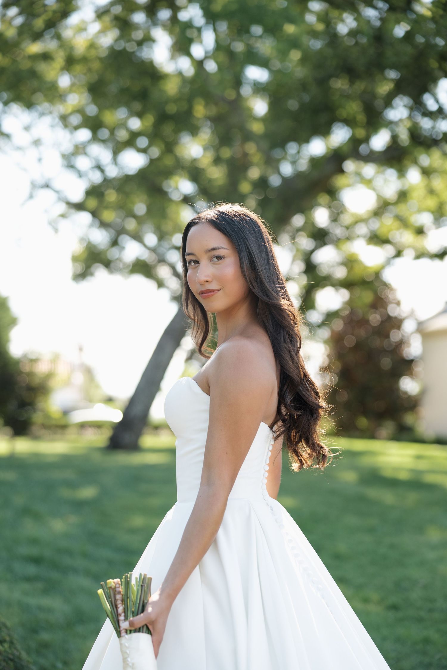 Bride in strapless white ball gown holding bouquet, posing outdoors on green lawn with large tree backdrop.